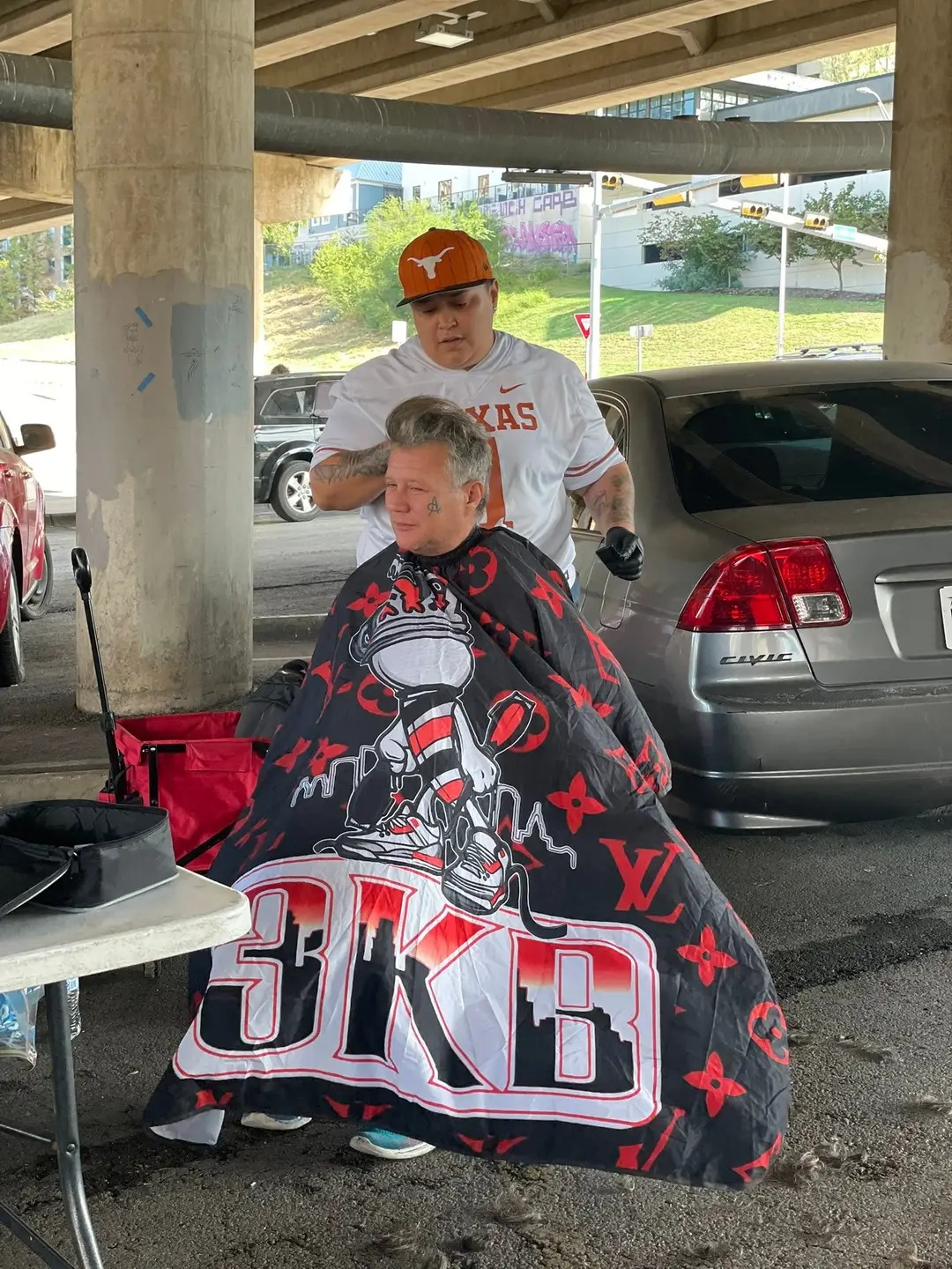 Person sitting in a chair with a barber cape featuring a basketball player and large text, under a parking structure.