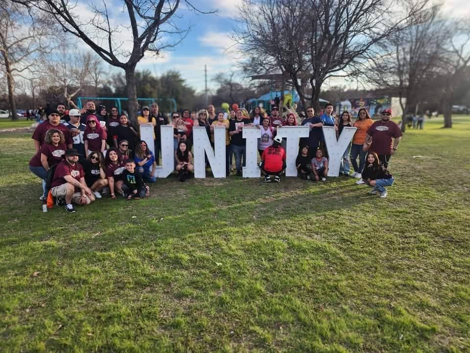 Group of people gathered outdoors with large letters spelling 'UNITY' in front of them, trees and a playground in the background.