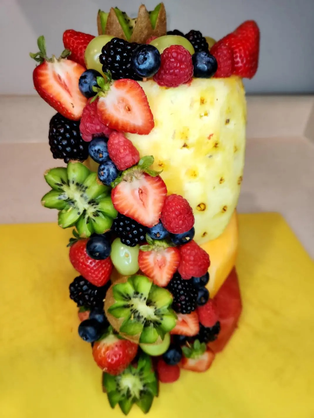 A tall glass filled with pineapple and surrounded by various fresh berries and sliced kiwi, with a yellow surface below.