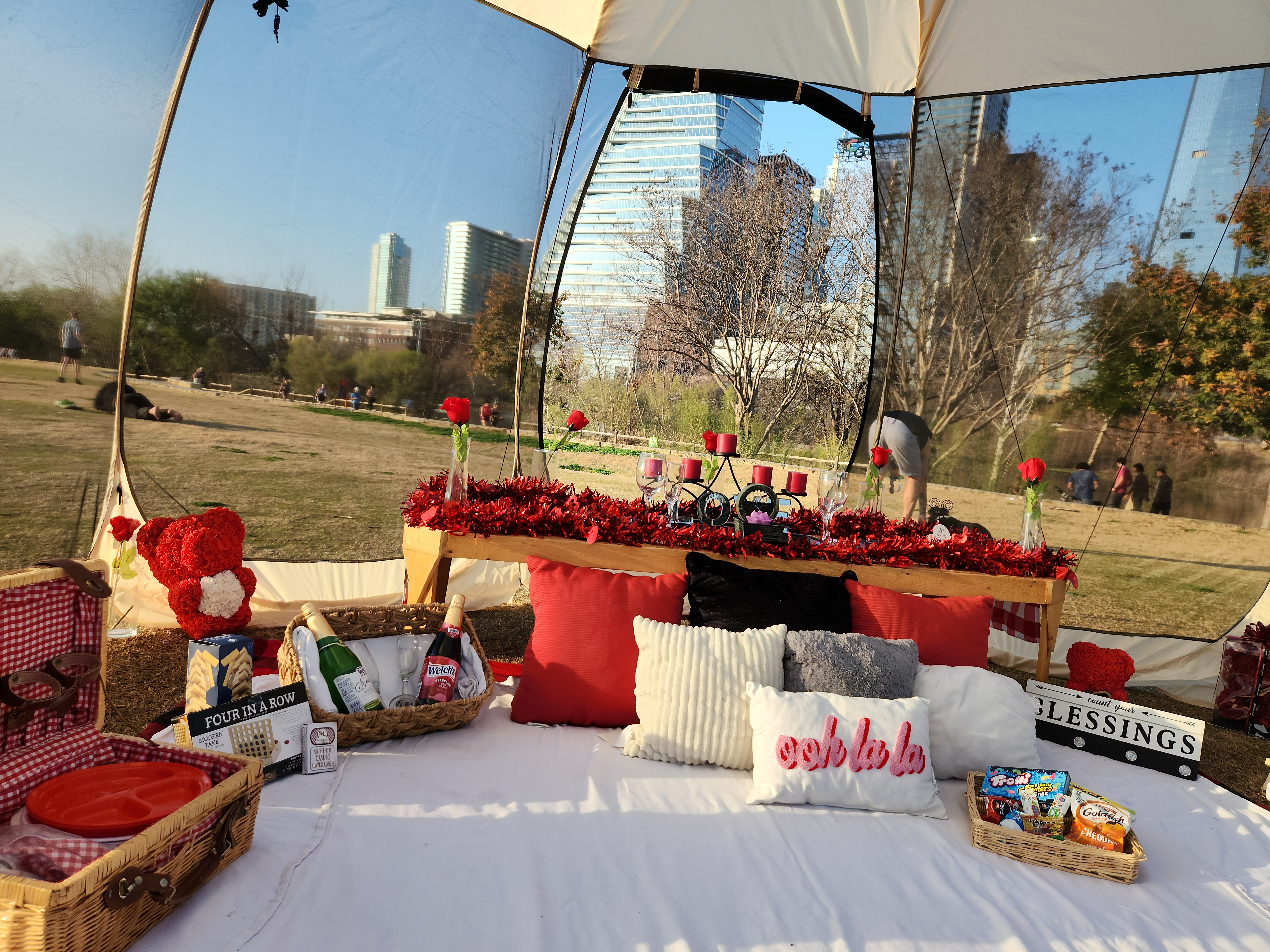 Outdoor setup with a table under a canopy, decorated with red flowers, candles, and gifts, in a park with buildings in the background.
