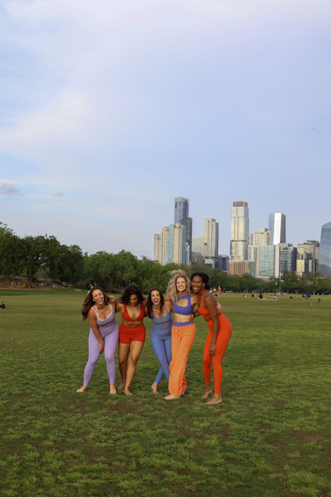 Four women stand together in a park with a city skyline in the background, smiling and posing for the photo.