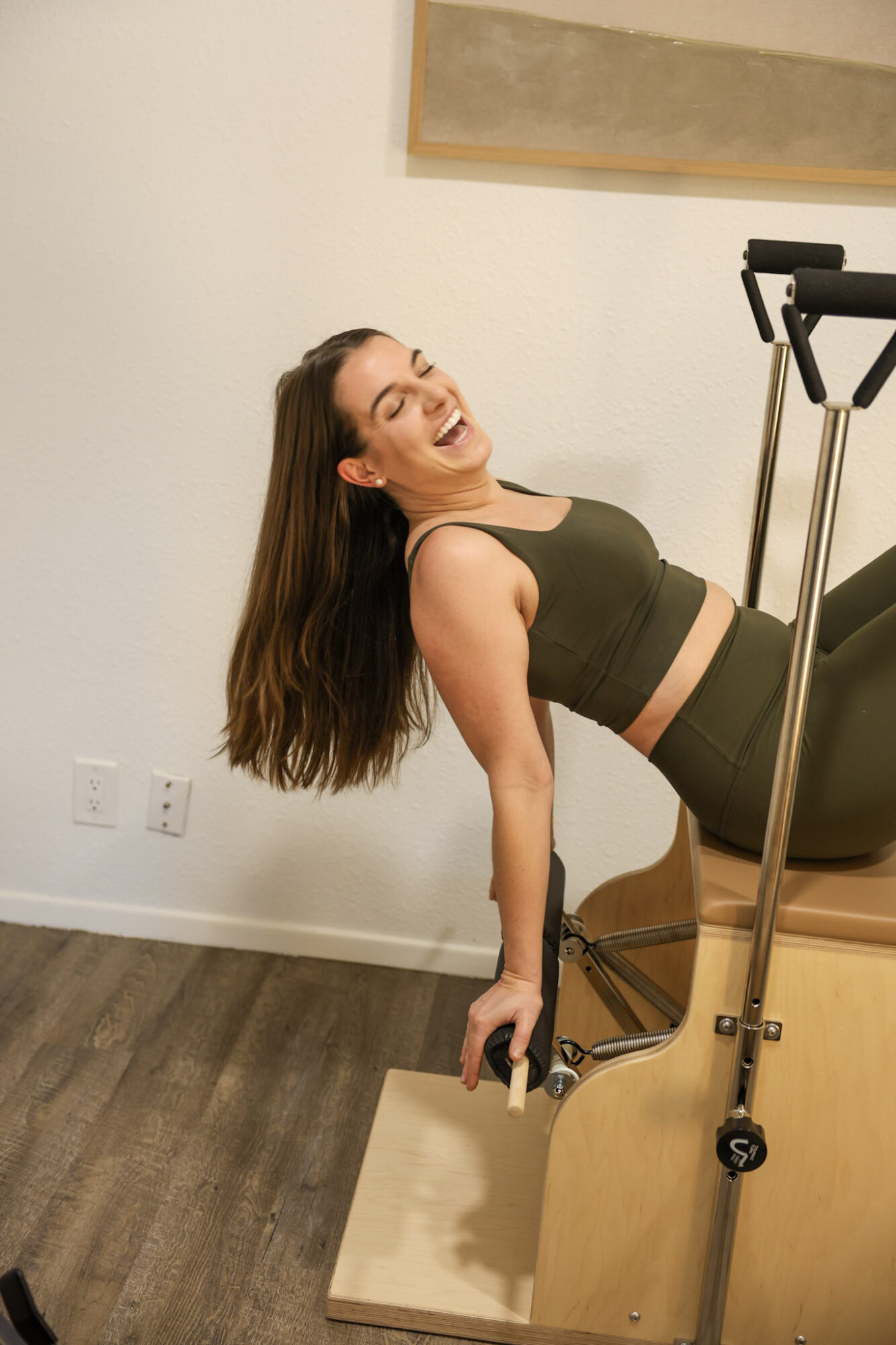 Woman smiling, leaning back on Pilates reformer machine, in workout attire, in a room with white walls and wooden floor.