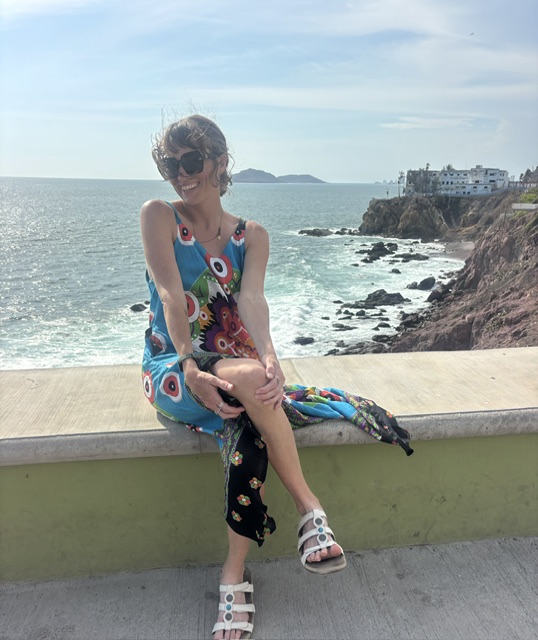 Woman sitting on a concrete ledge at the beach, smiling, wearing sunglasses, a colorful dress, and sandals, with ocean and cliffs in background.