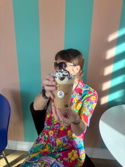 Person with sunglasses and colorful clothing holding a drink, taking a selfie in front of a striped background.