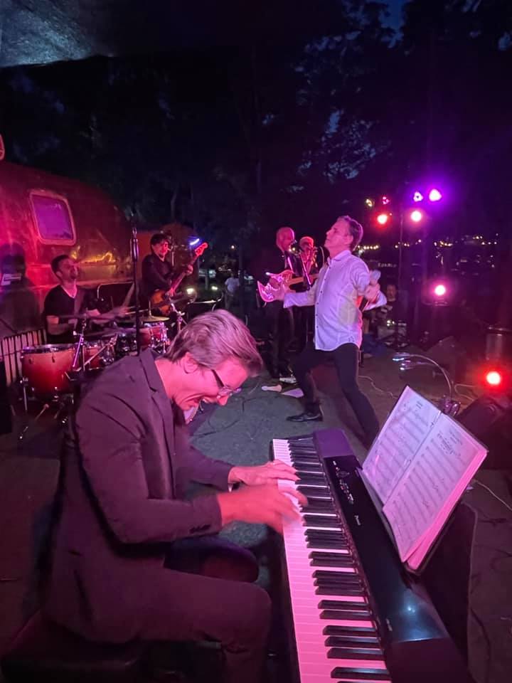 Musicians perform outdoors at night, with a pianist in the foreground and a band playing in the background, colorful stage lights.