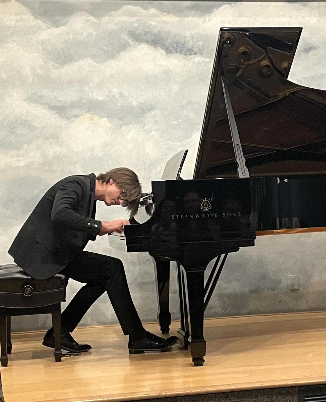 Person playing grand piano on stage with cloudy sky backdrop, leaning forward with head near the keys.