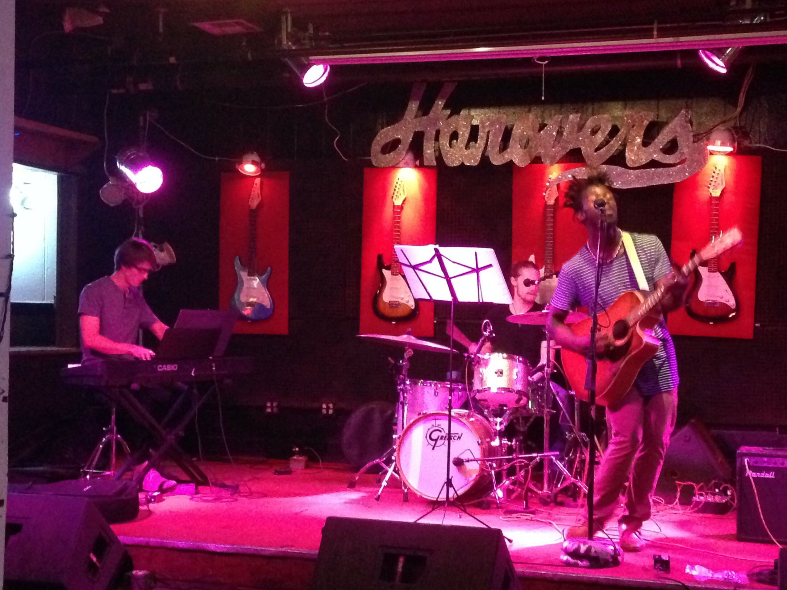 Three musicians perform on stage with guitars, keyboard, and drums, illuminated by pink and purple stage lights, with a sign in the background.
