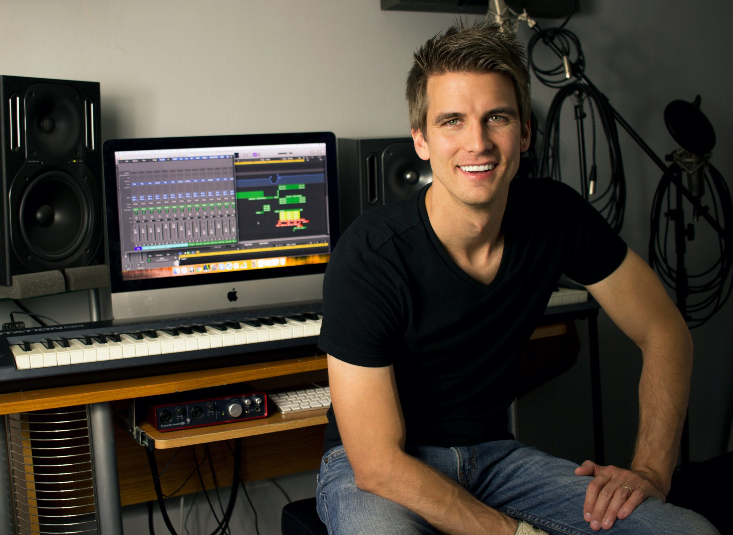 Young man sitting in a music studio with a computer displaying music production software, keyboard, and speakers.