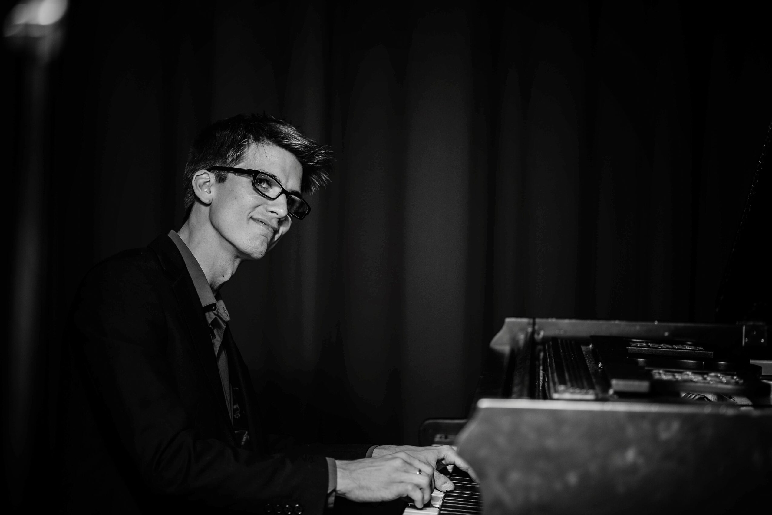 Person with glasses playing piano on stage with dark curtains in background.