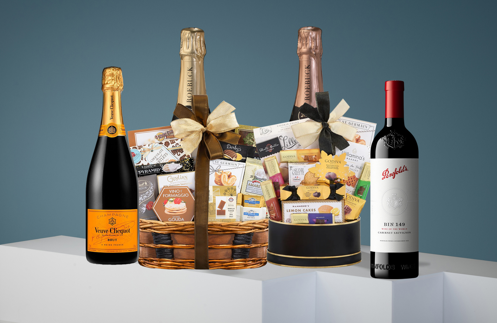 Gift basket with assorted snacks, two bottles of champagne, and a bottle of red wine on a white surface.