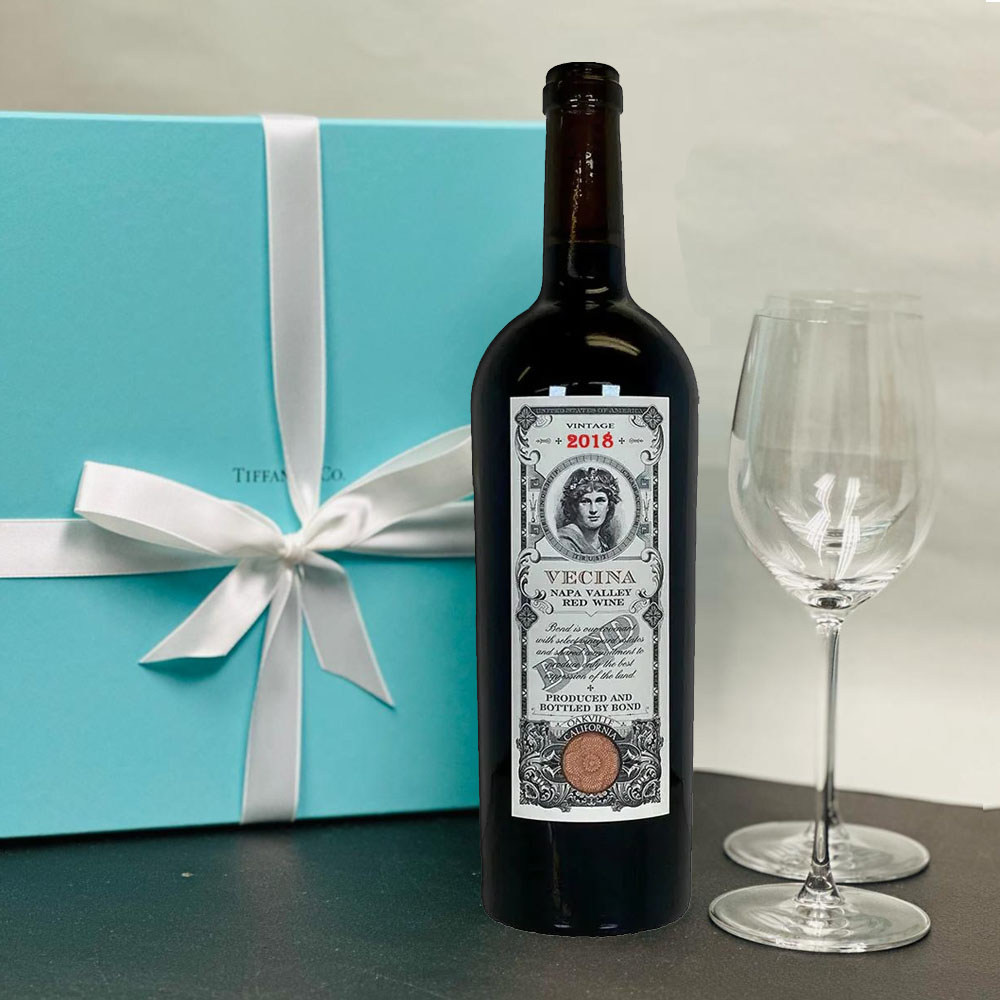 Bottle of wine with a label, empty wine glass, and a blue gift box with a white ribbon on a dark surface.