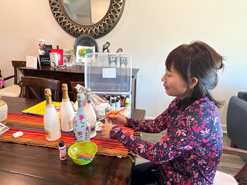 Woman with dark hair in a floral top sits at a table with bottles, a bowl, and a red cloth, smiling sideways.
