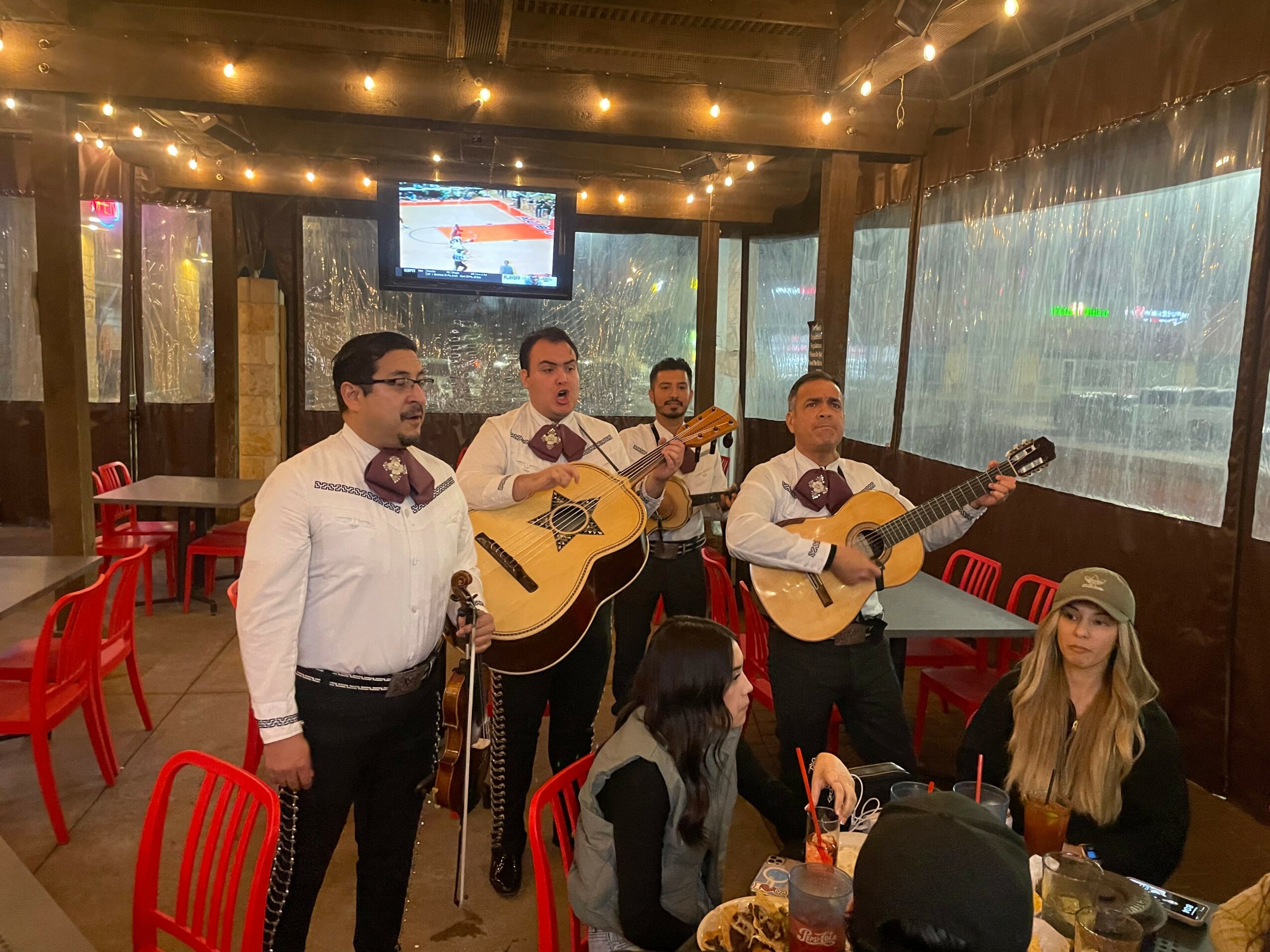 Four men playing guitars and singing, woman sitting at table, restaurant with red chairs and large windows, string lights overhead.
