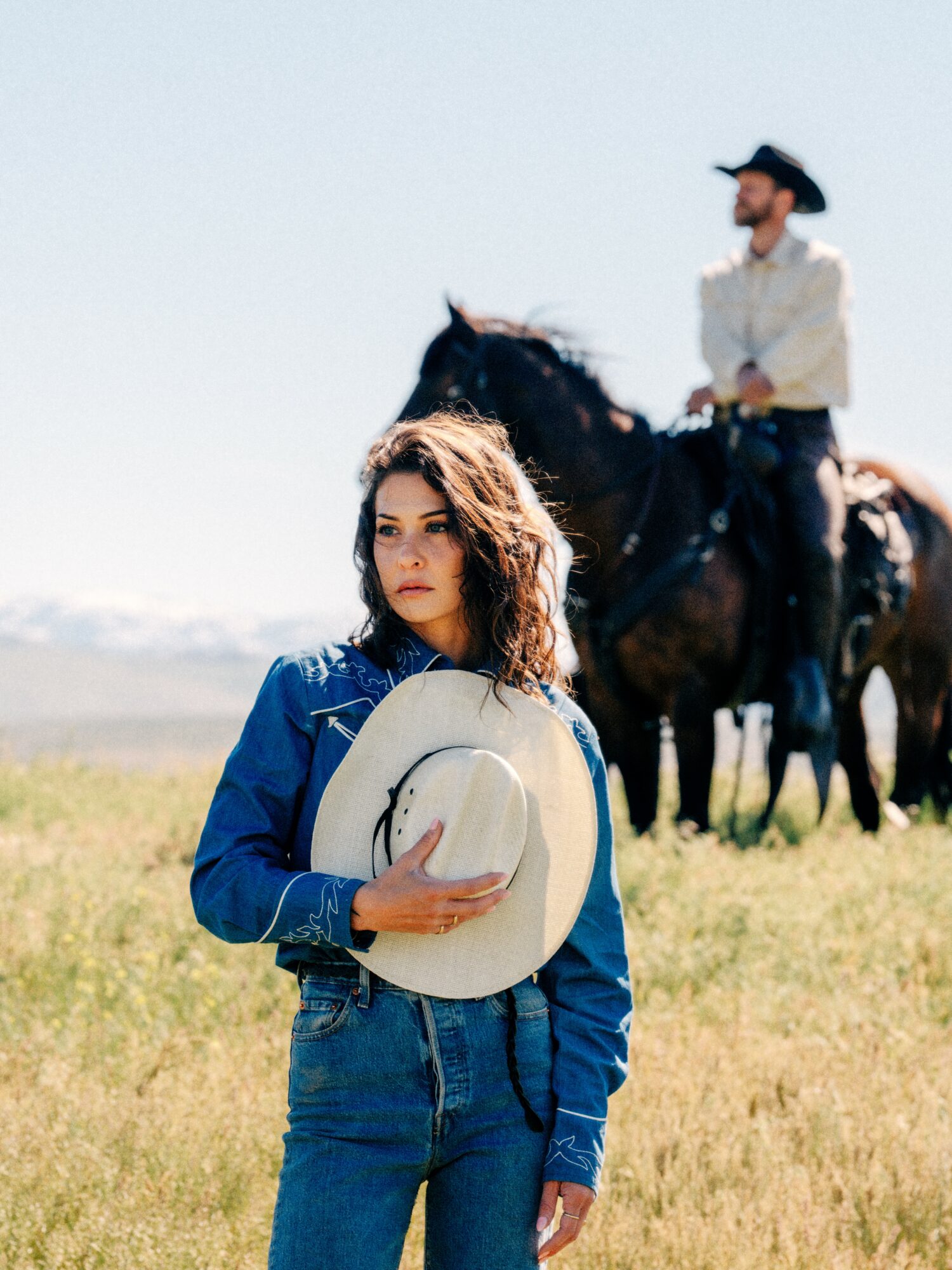 Young woman holding a hat, standing in a grassy field with a man on horseback in the background.