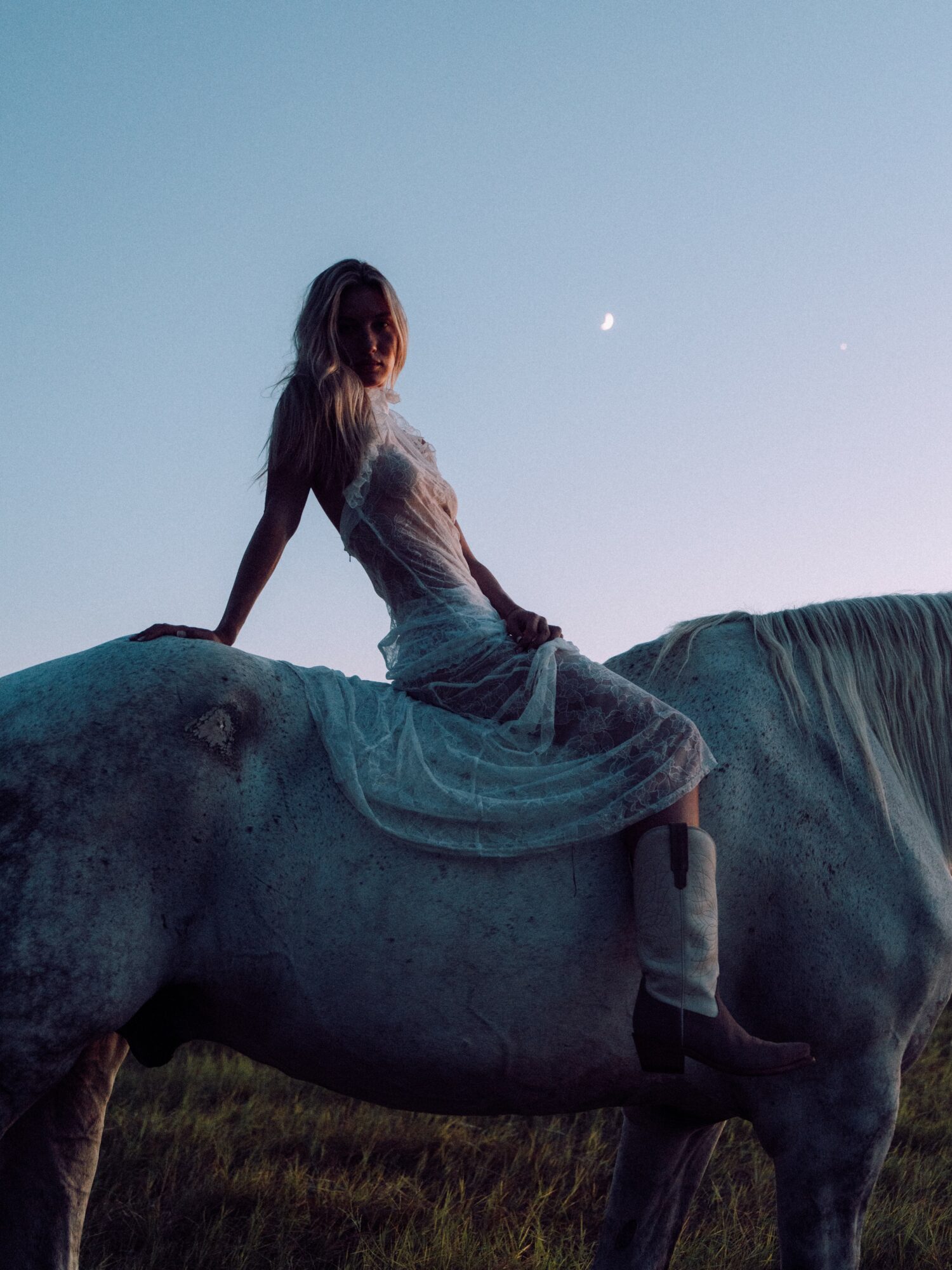 Person sitting on a large statue of a horse outdoors during dusk, with a visible moon in the sky.