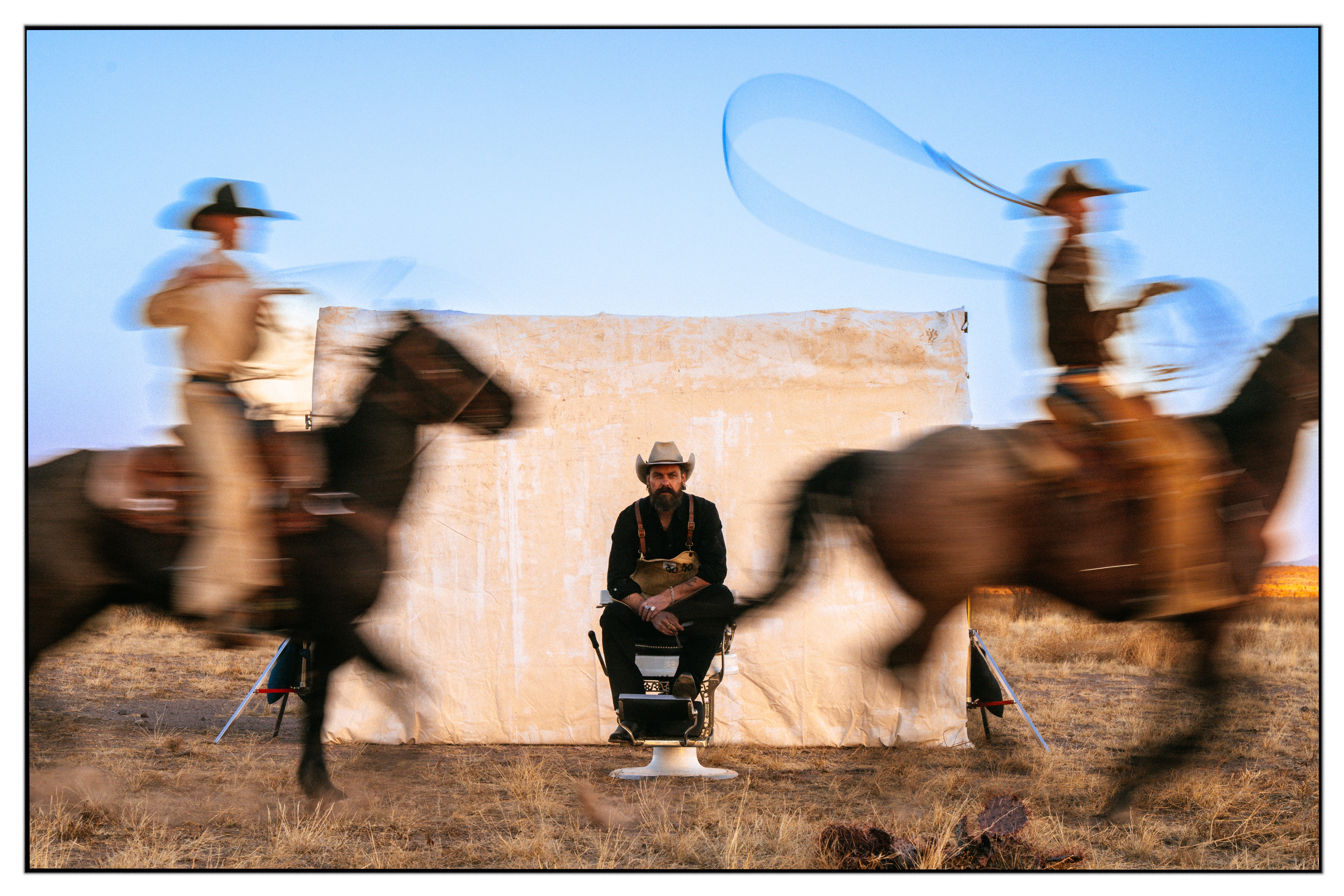 Person sitting on a chair between two moving horses with riders, against a wall, under a blue sky.