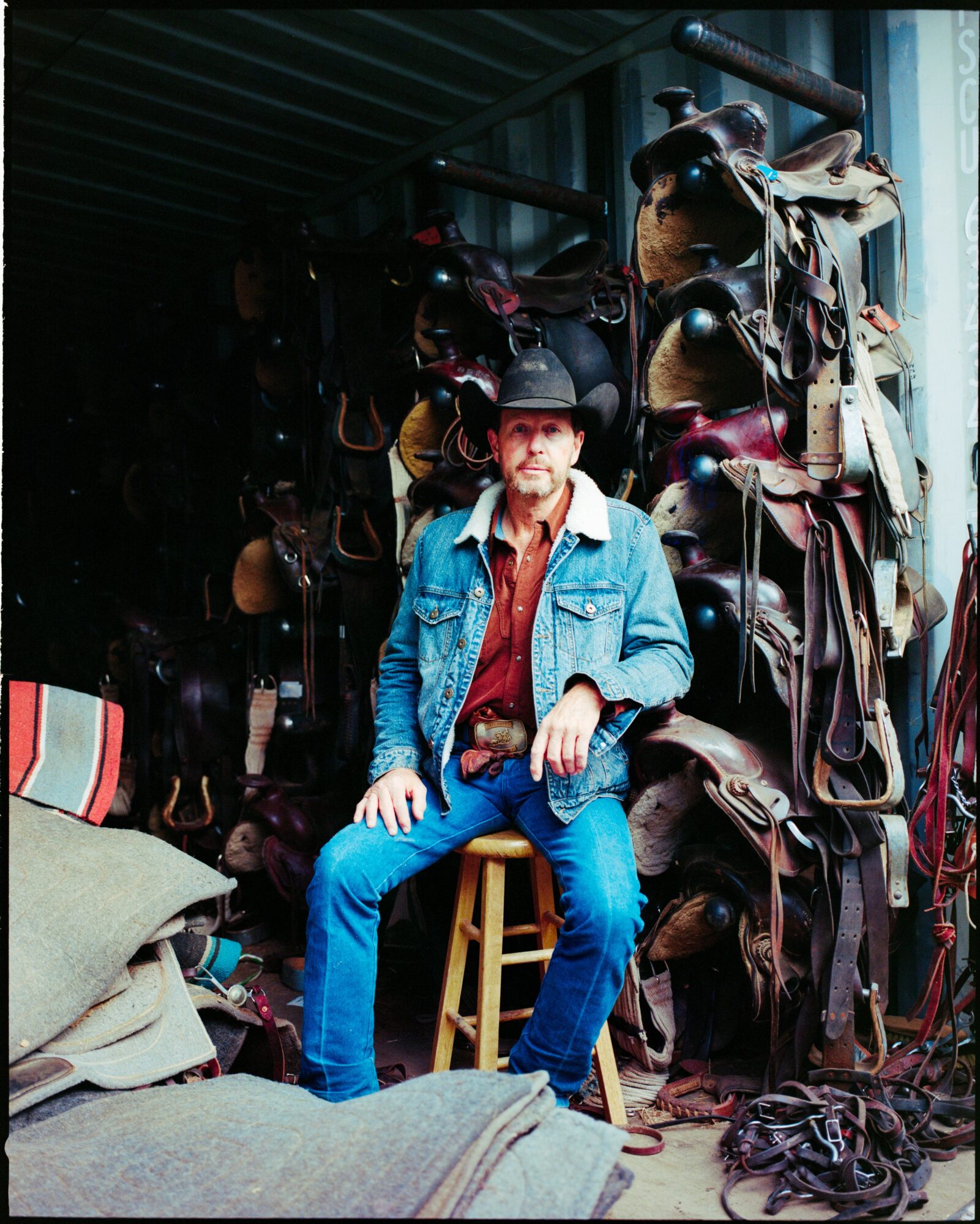 Man sitting on a stool in a storage area with helmets and gear hanging on the wall behind him.