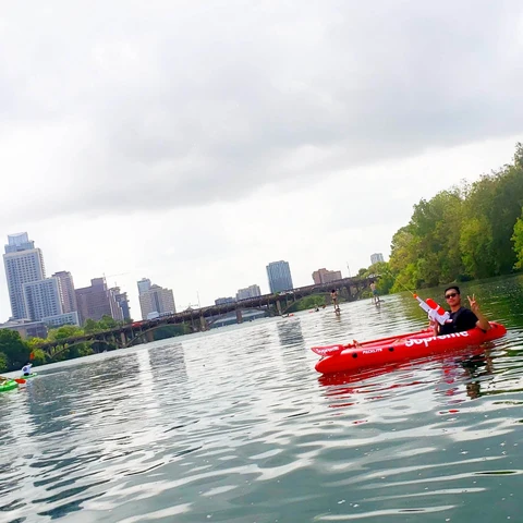 Person in a red kayak on a river with city skyline and trees in the background, overcast sky.