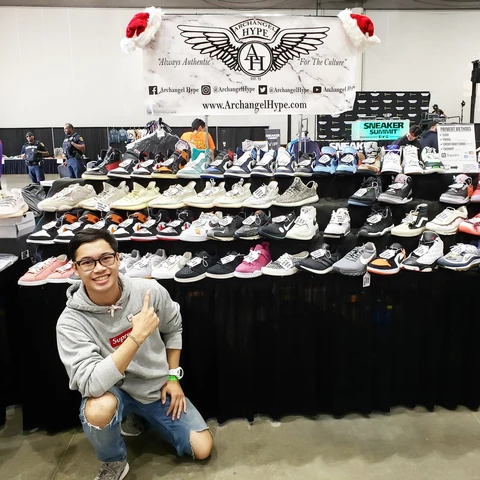 Person crouching in front of shoe display at event, smiling, wearing glasses, gray hoodie, and ripped jeans.