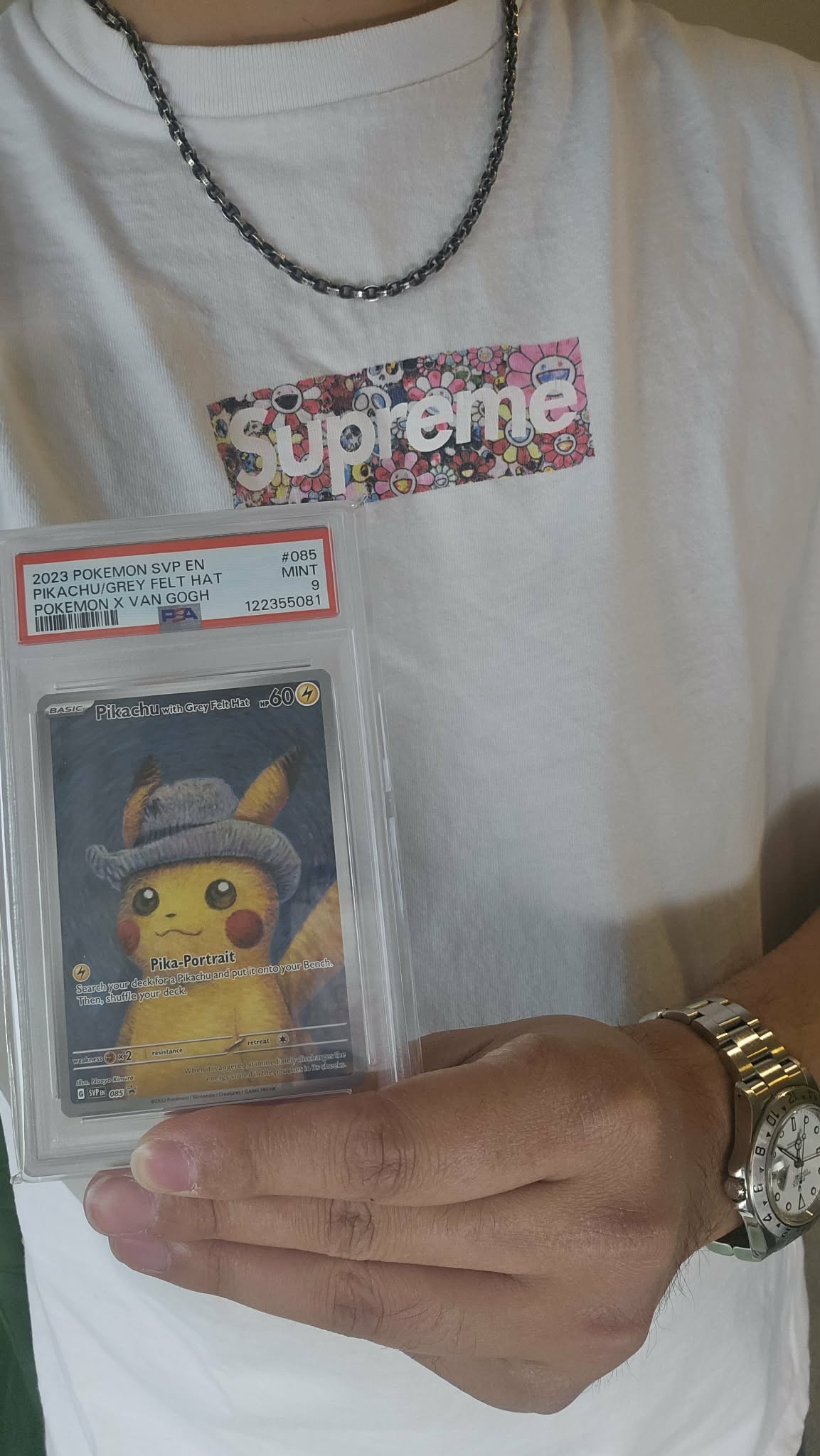 Person holding a graded Pokémon card in a protective case, wearing a white Supreme t-shirt, a silver chain, and a watch.