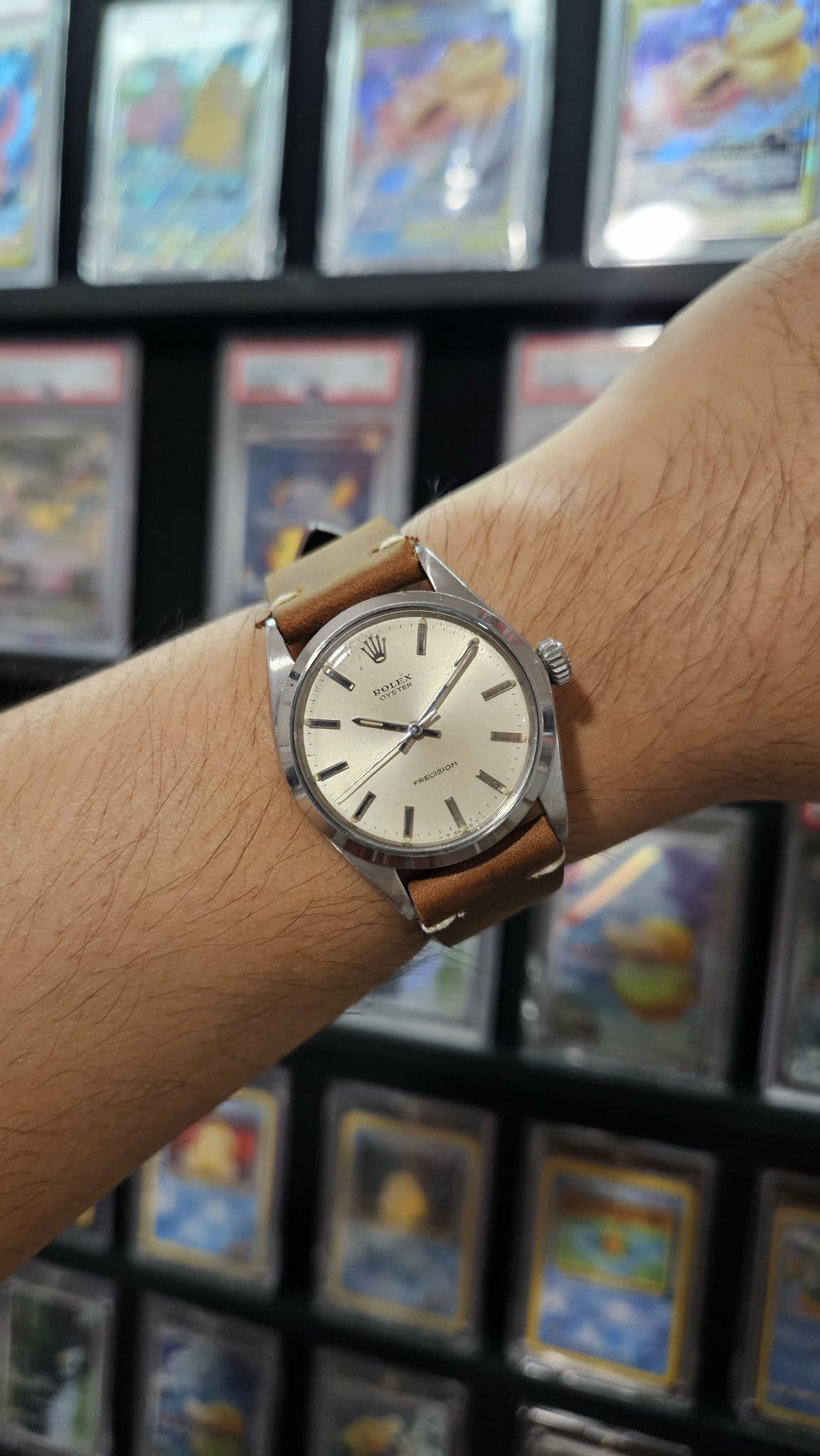 Person's wrist with a silver watch and brown leather strap, background shows blurred colorful items.