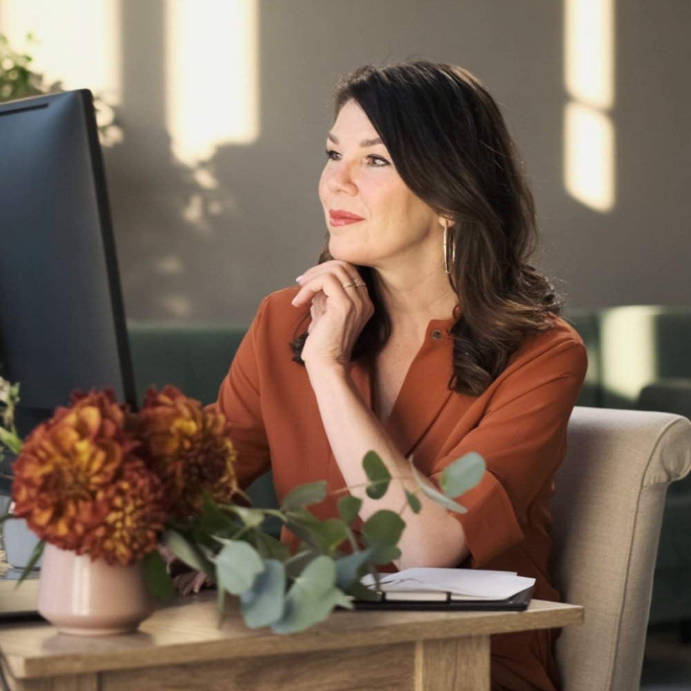 Woman with dark hair and earrings, wearing an orange top, sitting at a desk with flowers and a notebook, looking at a computer screen.