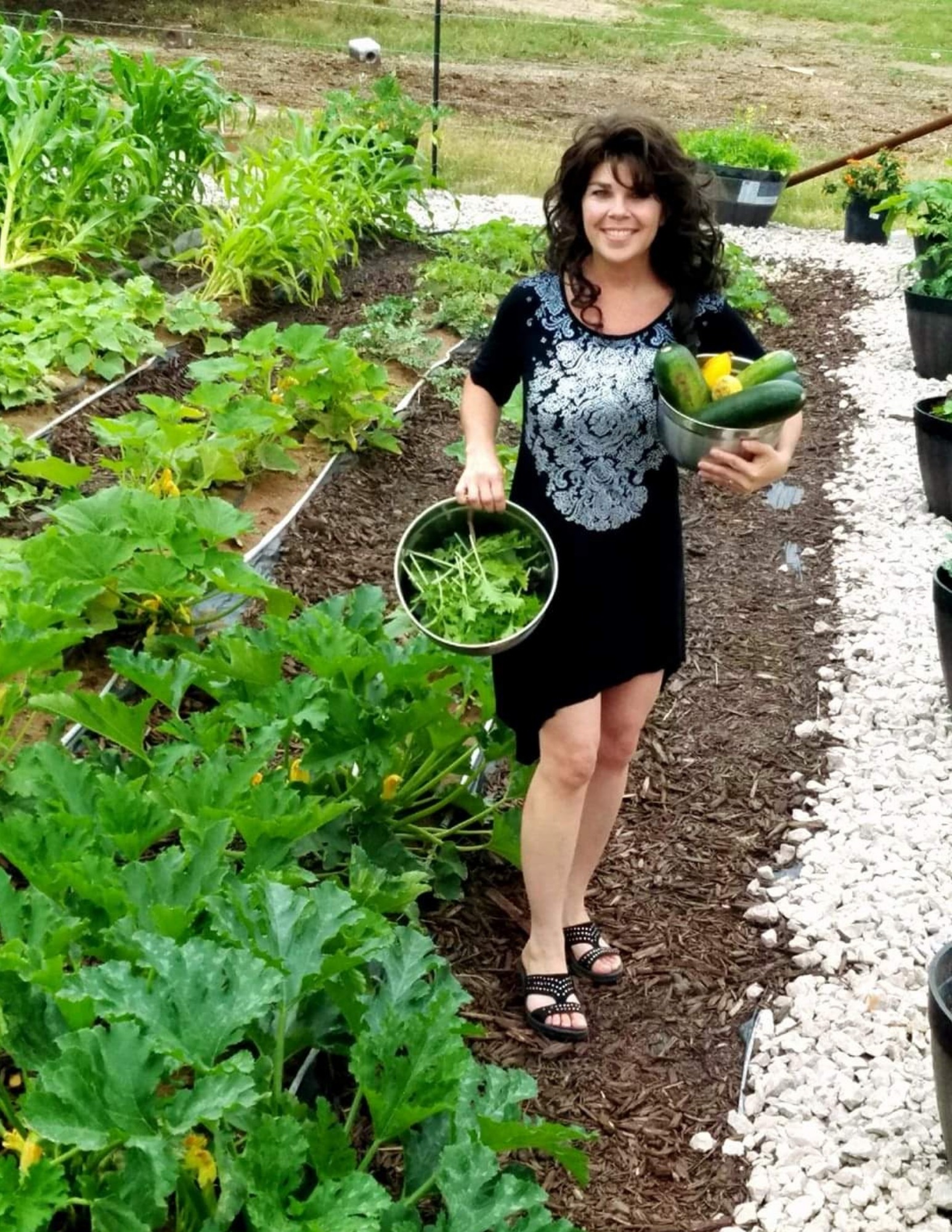 Woman with curly hair holding a basket of vegetables and a small bucket in a garden, smiling, surrounded by plants.