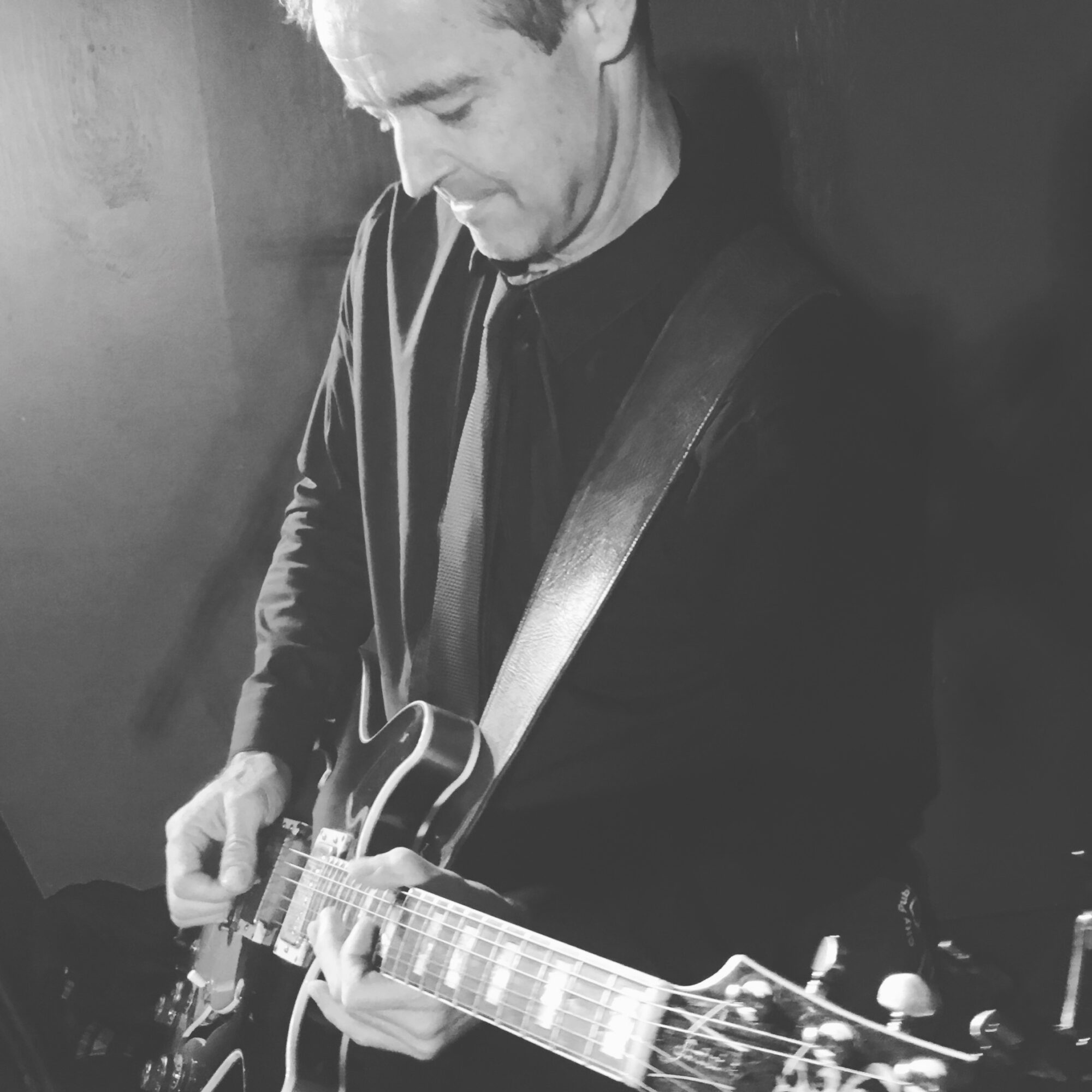 Man playing an electric guitar, looking down, in black and white, wearing a dark jacket and shirt.