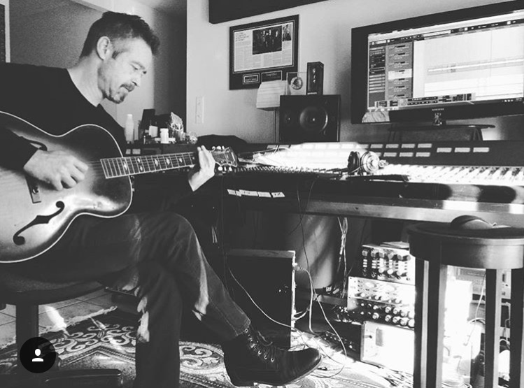 Person playing guitar in a music studio with computer and audio equipment, black and white image.