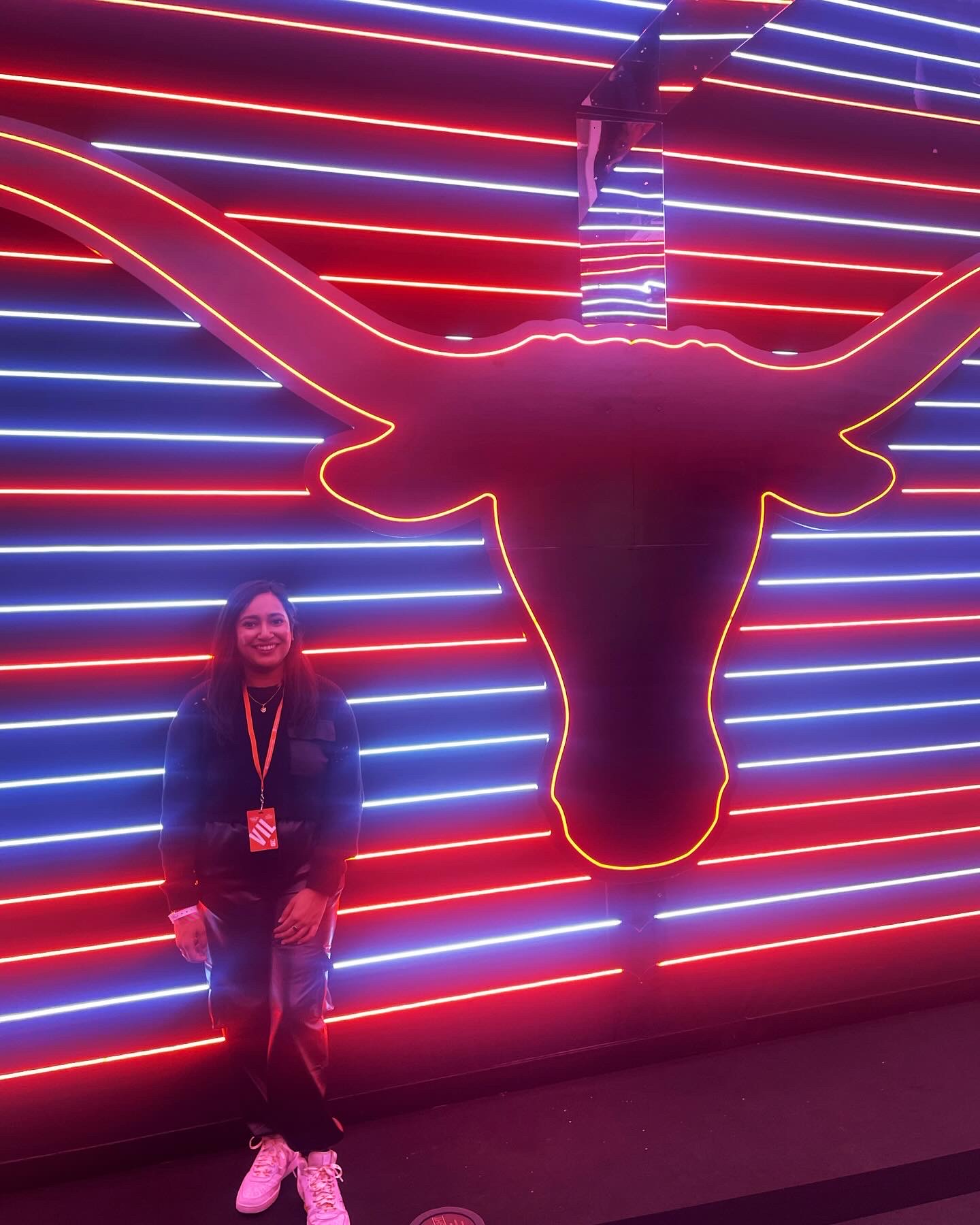 Person standing in front of a neon Texas Longhorns logo with red and blue lights, smiling, wearing a jacket and lanyard.