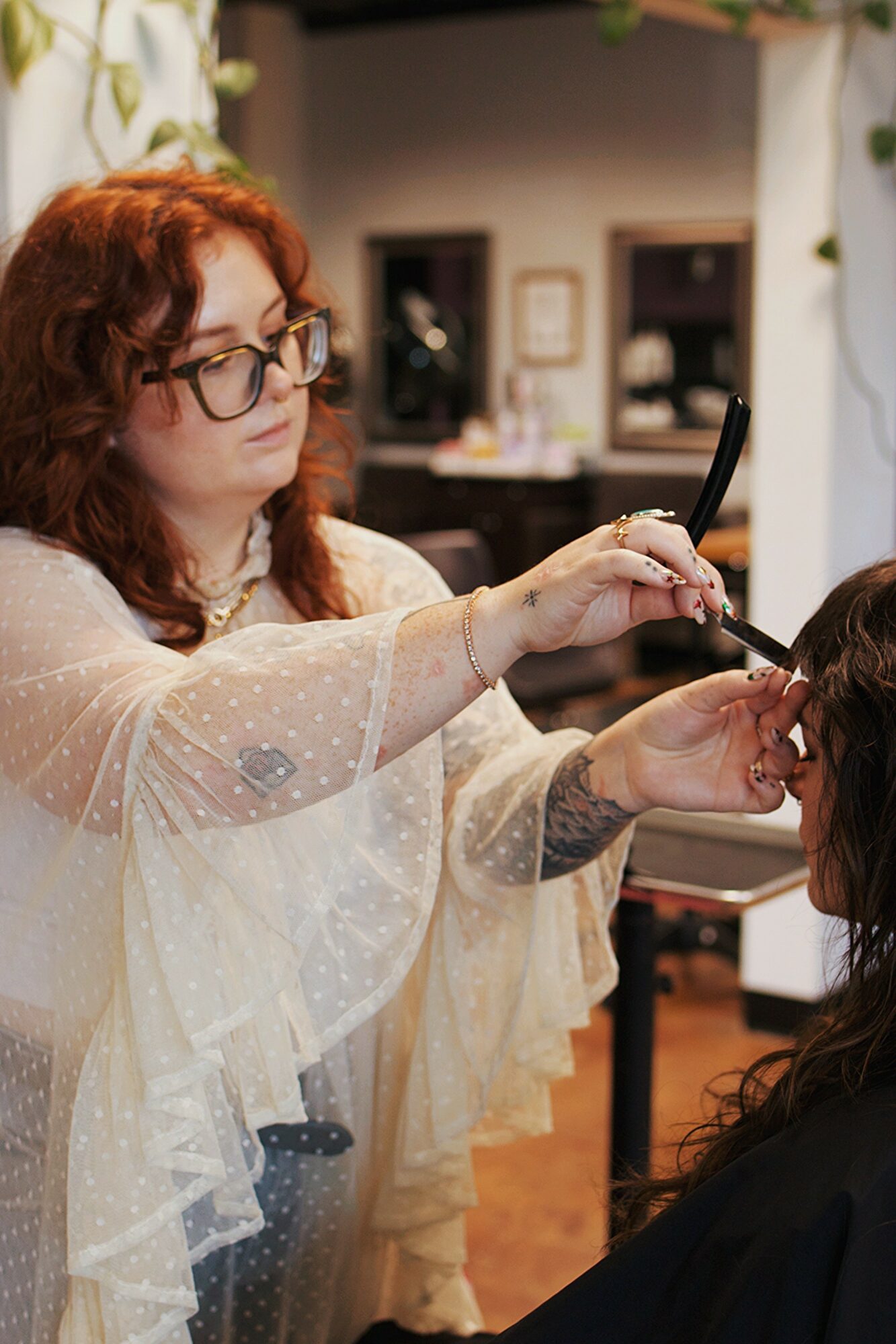 A woman with curly red hair and glasses is applying makeup to another person in a salon, with shelves and mirrors in the background.