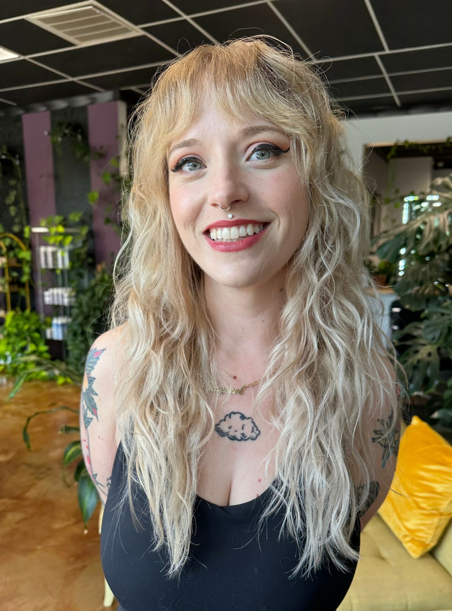 Young woman with long, wavy blonde hair smiling indoors, wearing a black top, with plants and furniture in background.