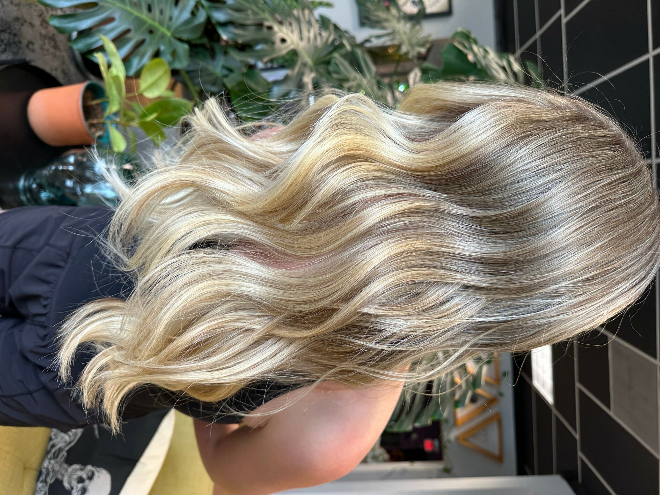 Side view of a person with long, wavy, blonde hair, in a setting with plants and black tiled wall.