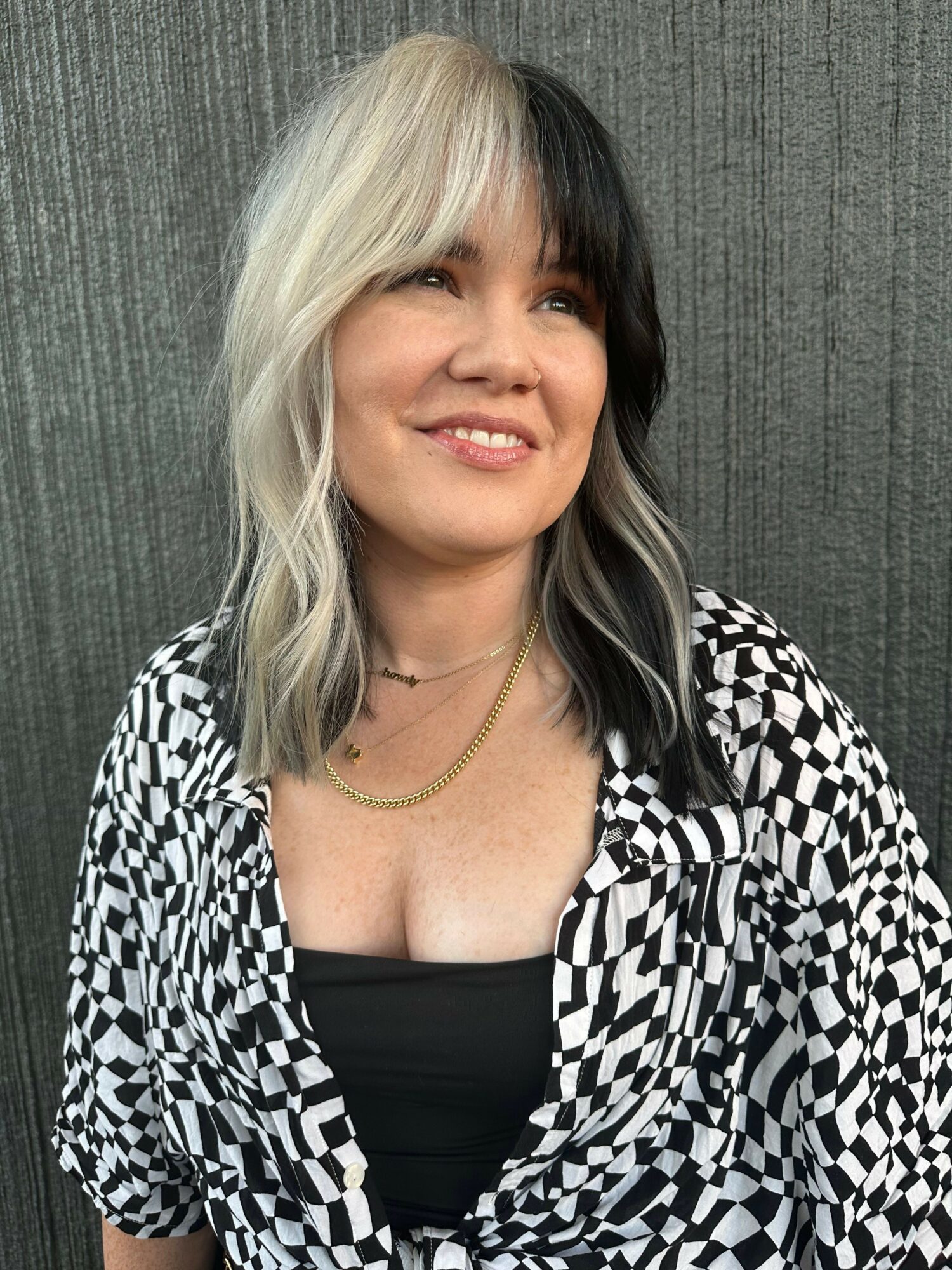 Woman with blonde hair smiling, wearing a checkered jacket and black top, against a textured gray background.