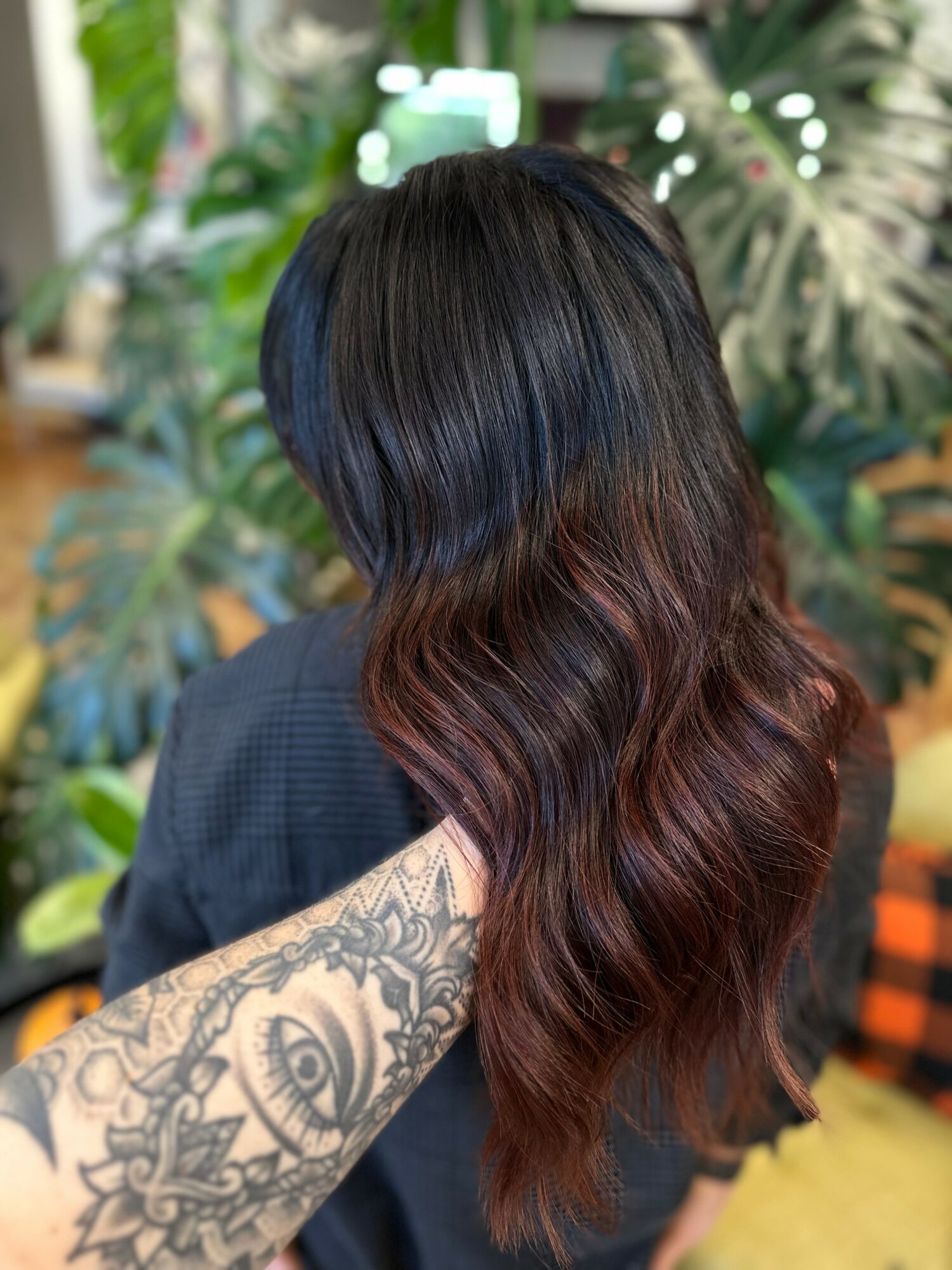 Person with long, wavy hair transitioning from black to reddish-brown, holding their hair with a tattooed arm, in a green indoor setting.
