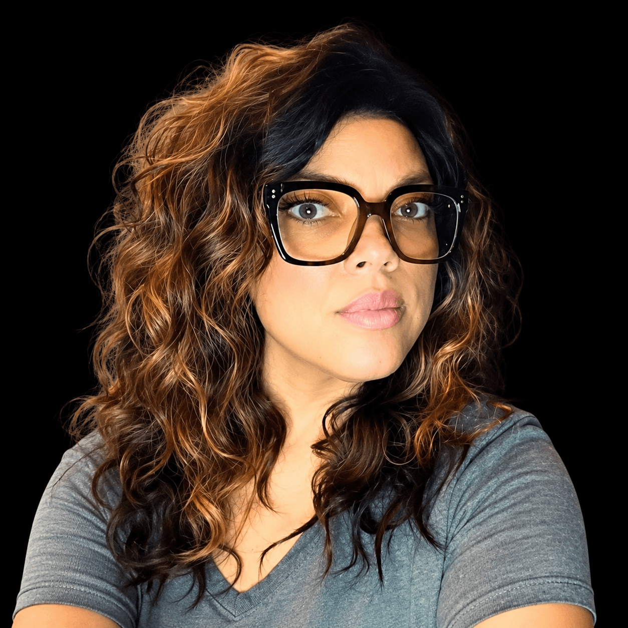 Woman with curly hair, glasses, and a gray shirt looks to the side against a black background.