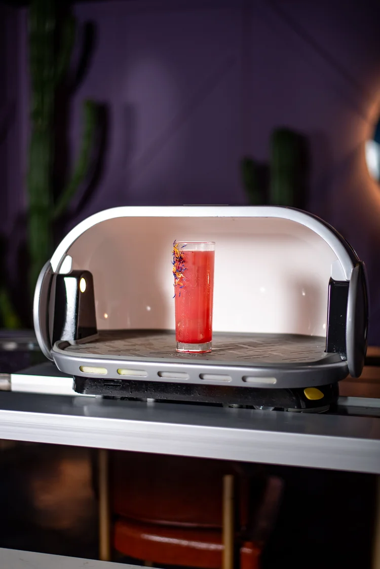 A red drink in a tall glass on a device with a transparent shield, in a dimly lit room.