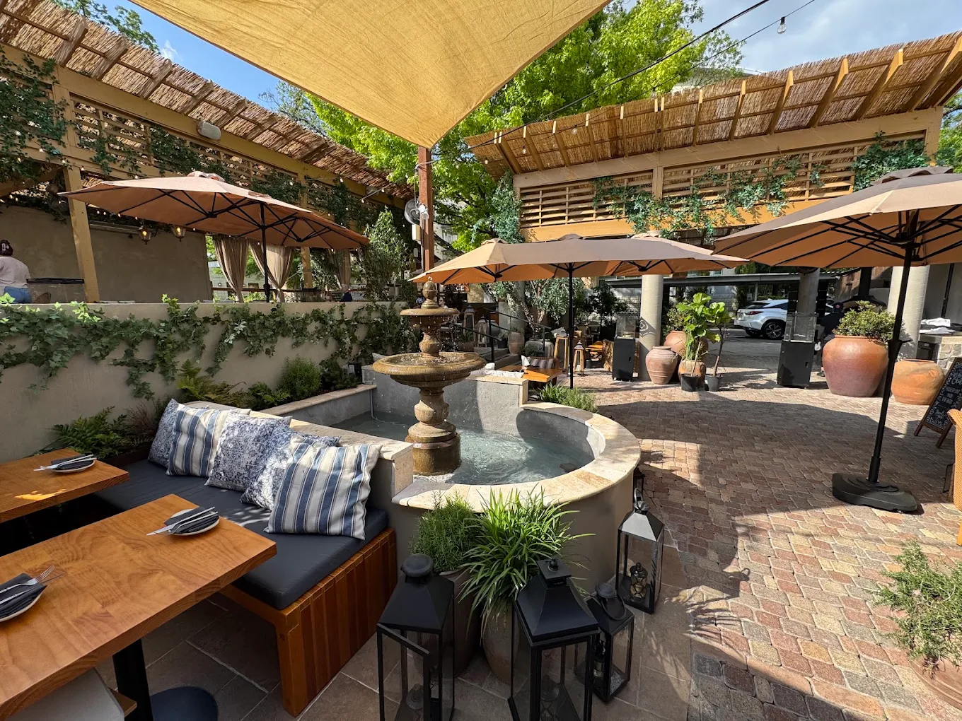 Outdoor patio with tables, umbrellas, and a fountain, surrounded by greenery and shaded by a canopy.