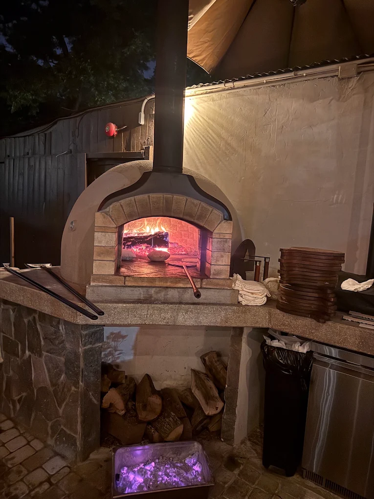 Outdoor pizza oven with a fire inside, surrounded by stone and wood, under a canopy, with stacked plates nearby.