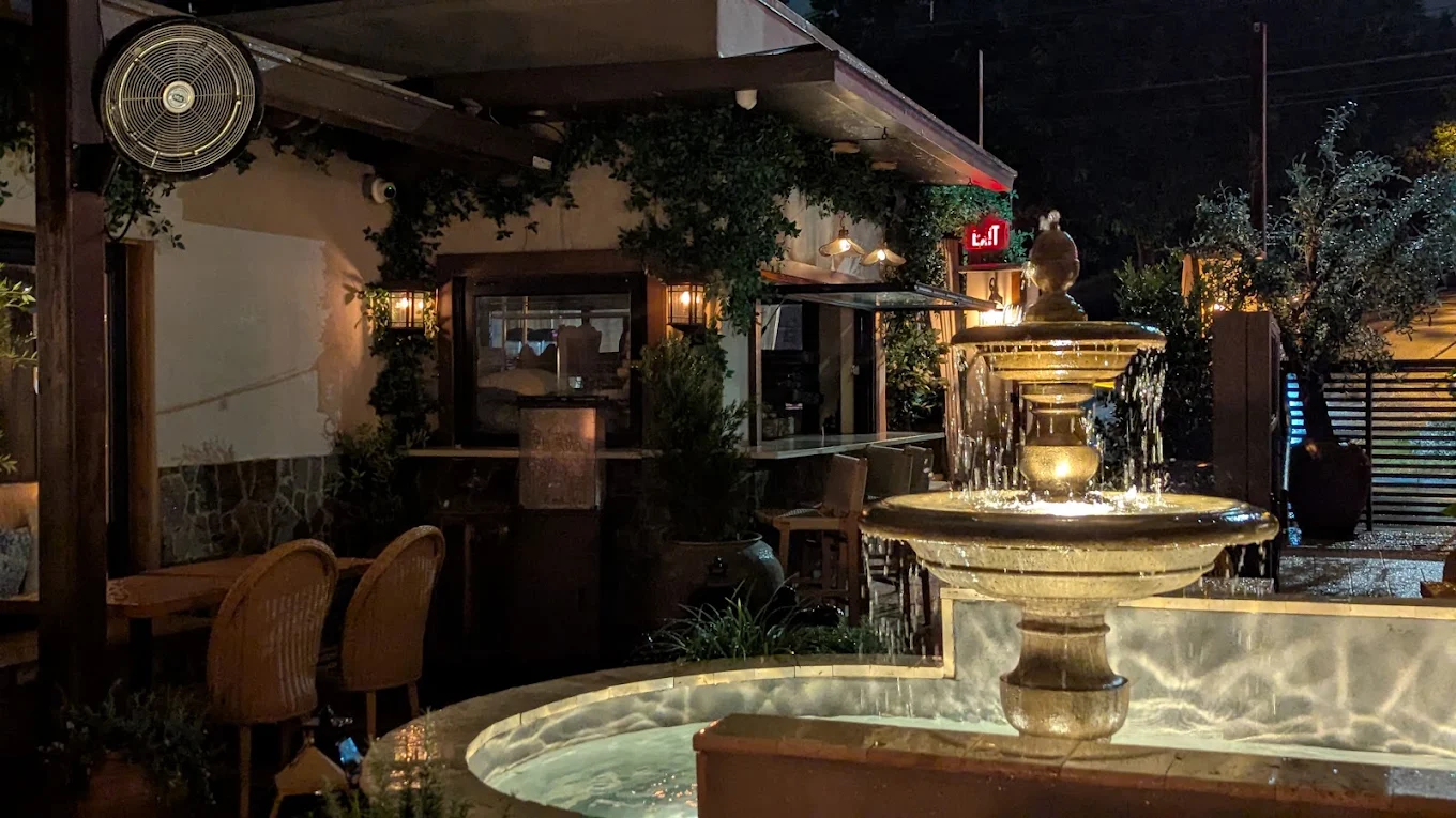 Nighttime outdoor scene with a fountain, chairs, and a building with warm lighting, trees, and a person in the background.
