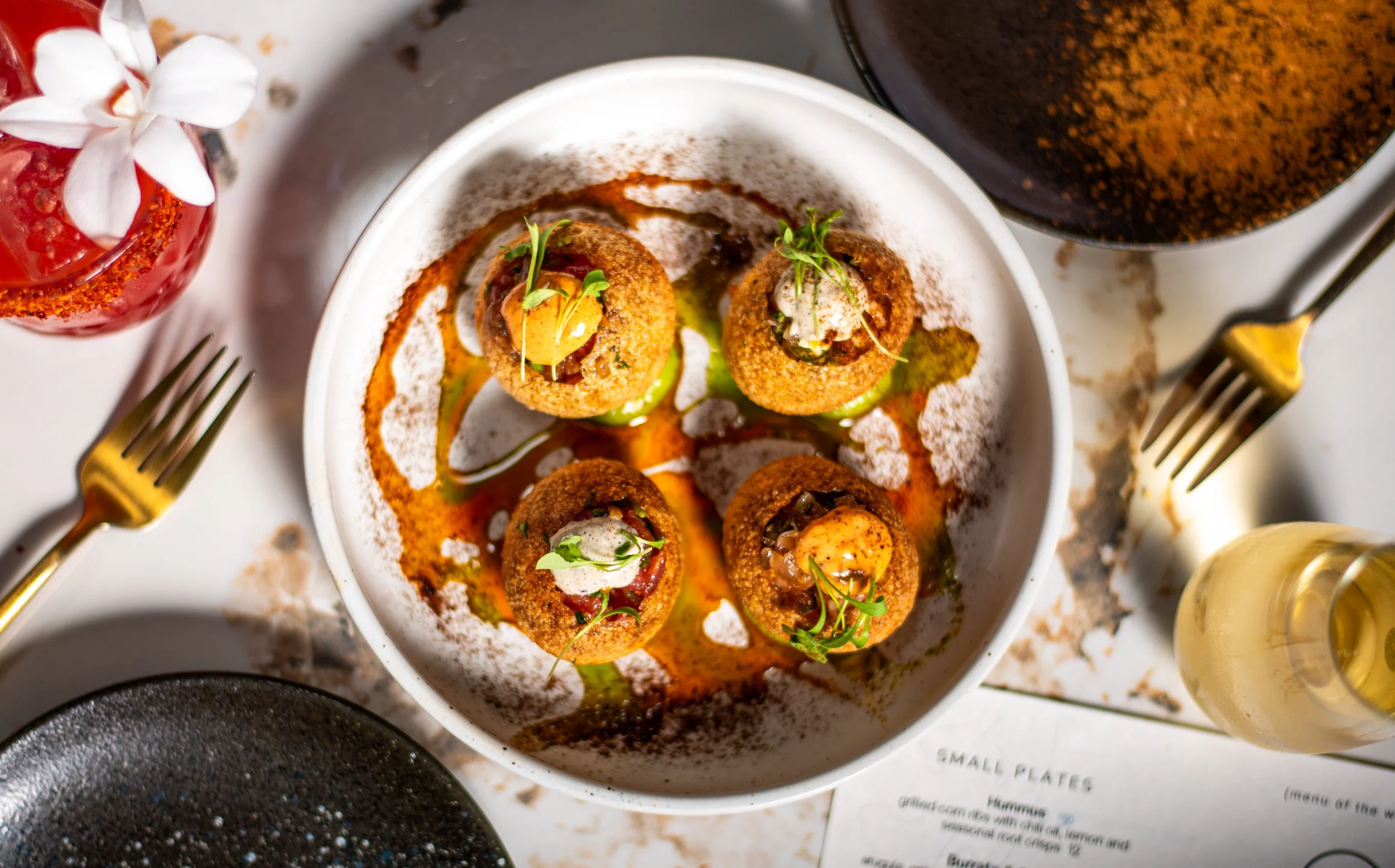 Four stuffed appetizers on a white plate with sauce, surrounded by gold utensils and a glass of white wine.