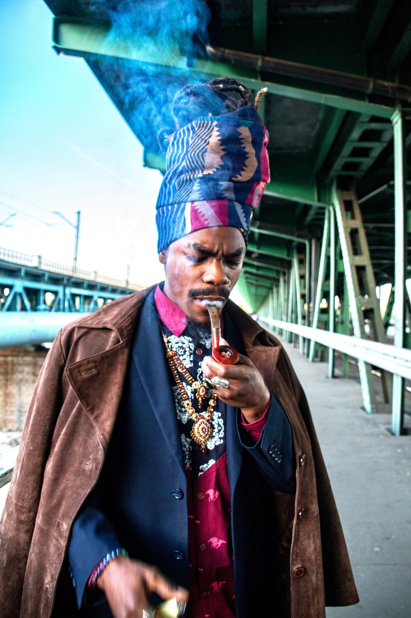 Man with colorful headwrap and jewelry smokes a pipe under a bridge, wearing a brown coat and dark suit.