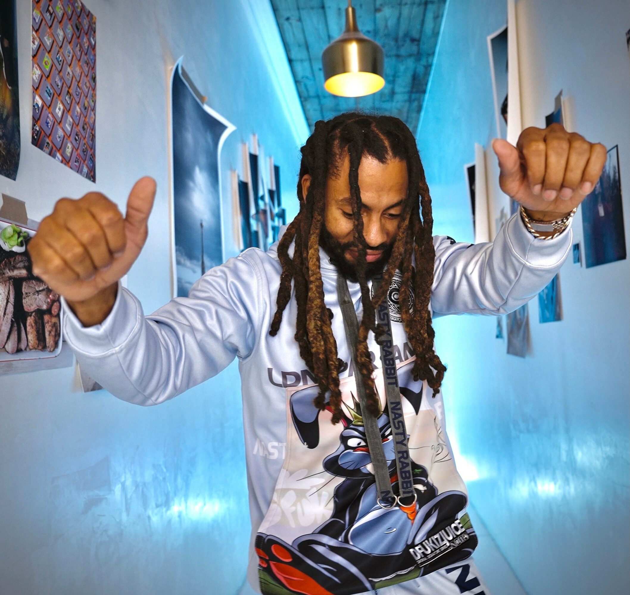 Person with dreadlocks and white jacket dancing in a hallway with blue lighting, arms raised, colorful artwork on walls.