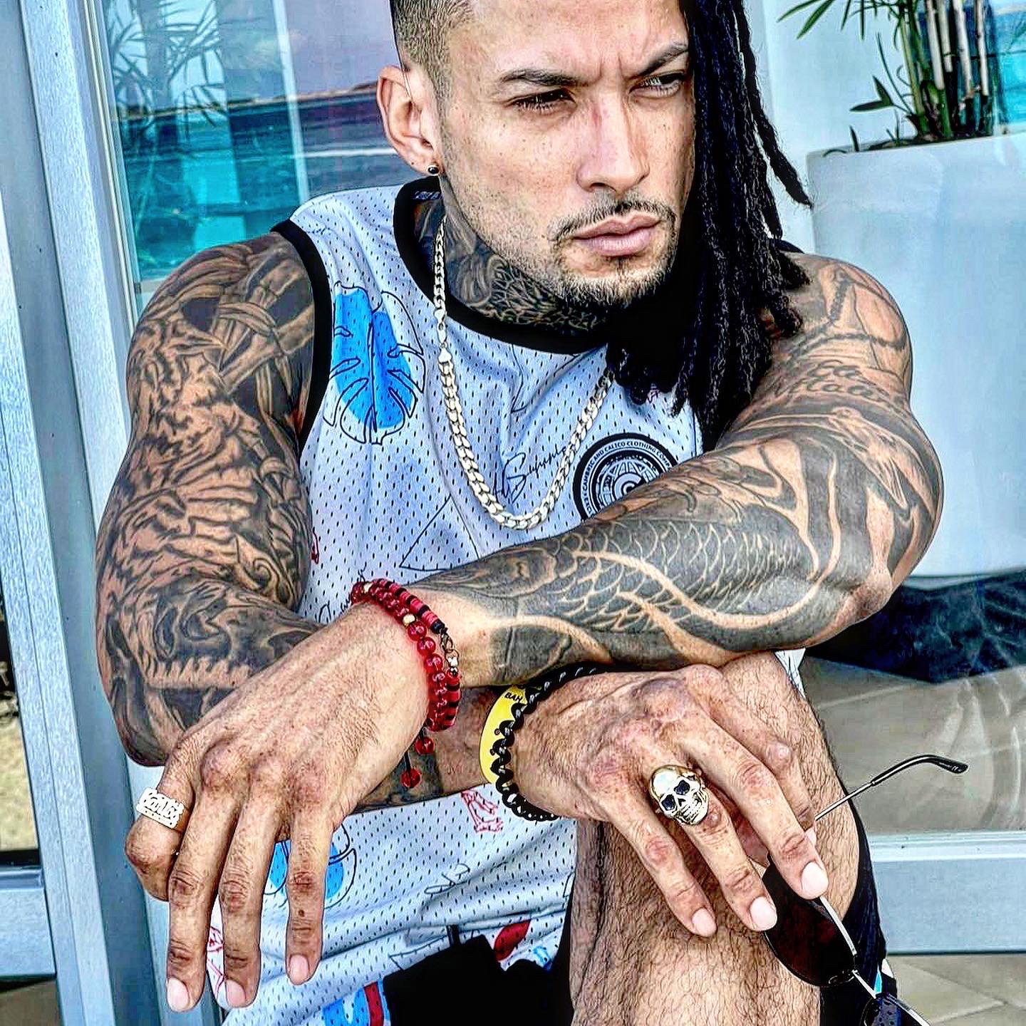 Tattooed man with dreadlocks, wearing a sleeveless shirt, jewelry, and sunglasses, sitting outdoors with arms crossed.