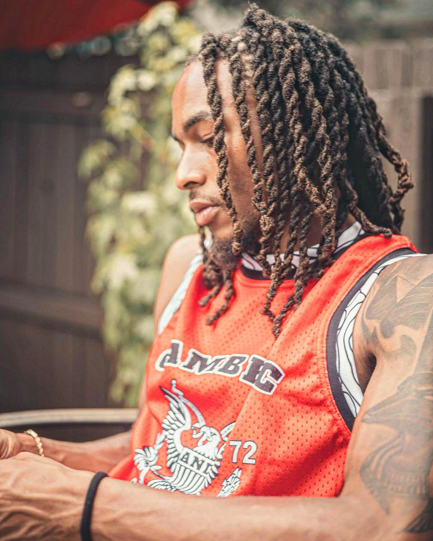 Man with dreadlocks wearing a red sports jersey outdoors, looking to the side, with a blurred background of trees and a fence.