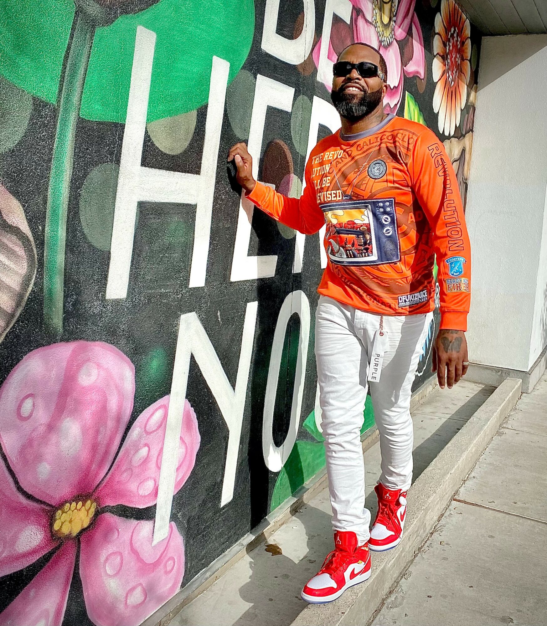 Man with sunglasses and beard in orange sweatshirt and white pants standing against colorful mural wall.