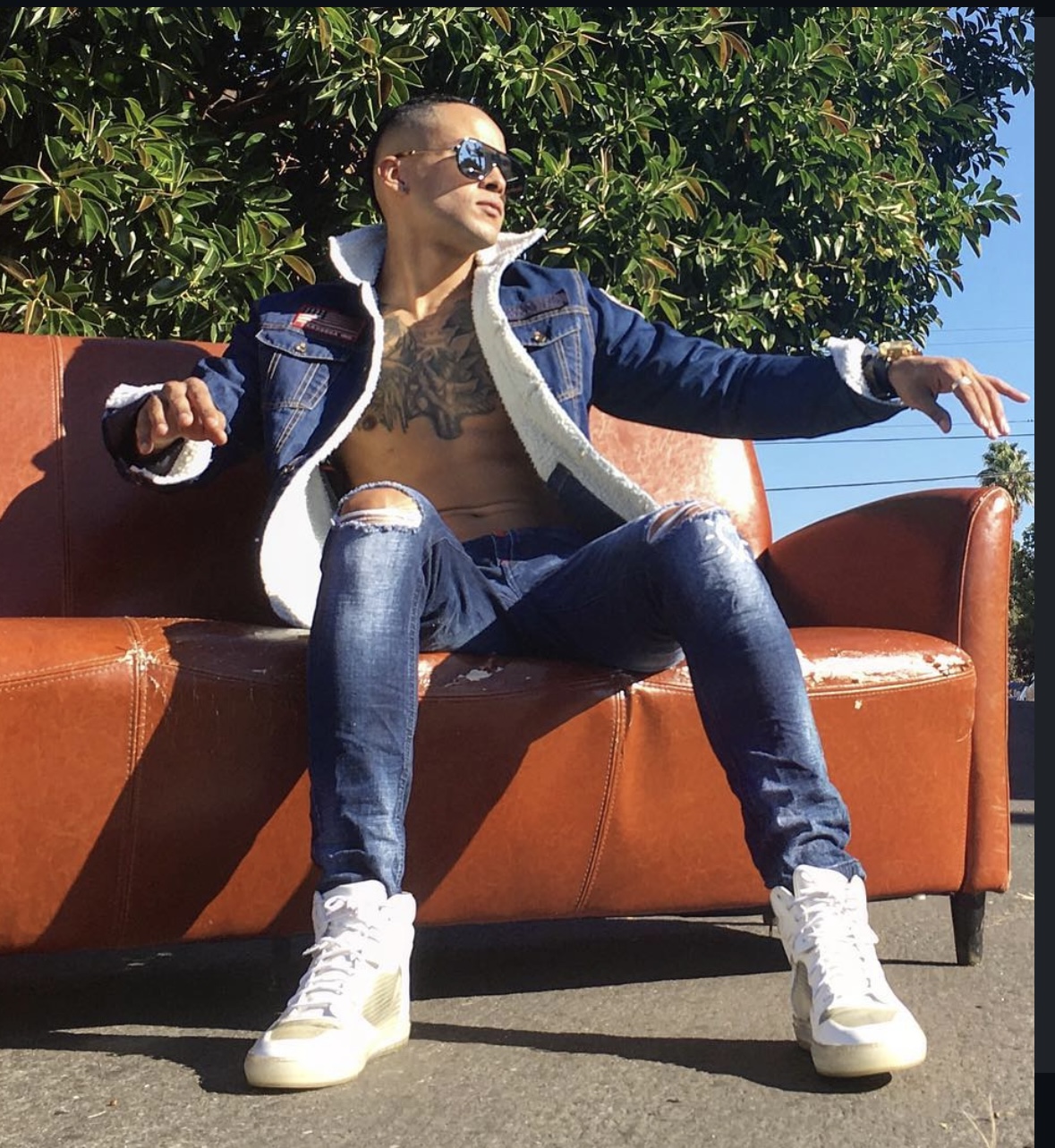 Person sitting on a brown outdoor sofa, wearing sunglasses, denim jacket, ripped jeans, and white sneakers, with greenery and blue sky behind.