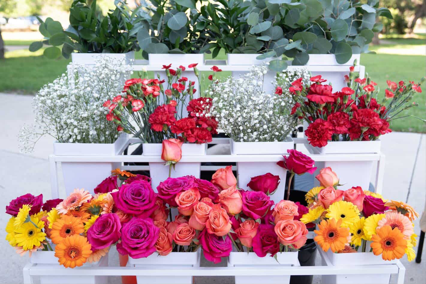 Multiple colorful flowers including roses, daisies, and other blooms arranged in pots and containers outdoors.
