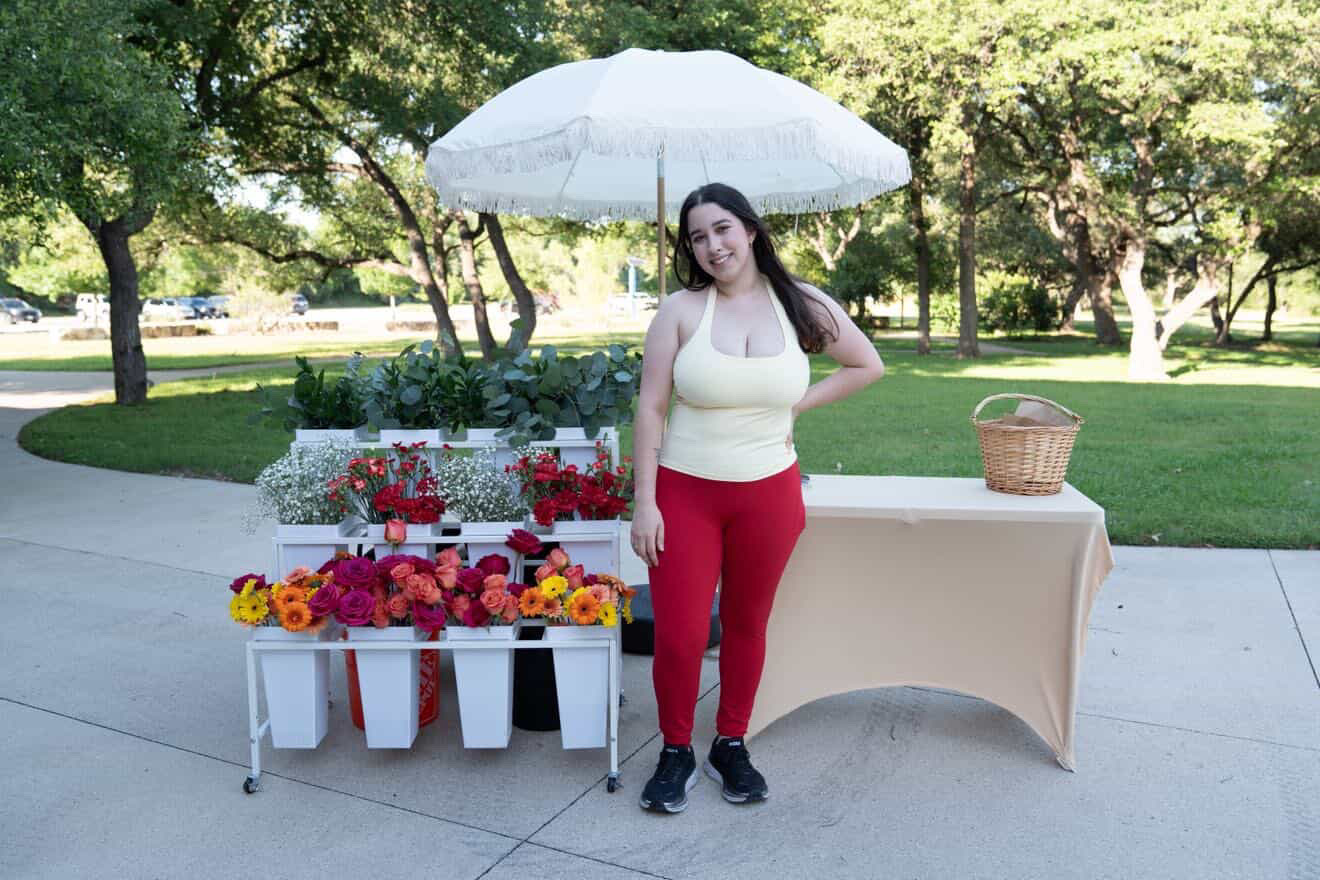 Woman standing next to flower display and table outdoors, holding an umbrella, with trees and grass in background.