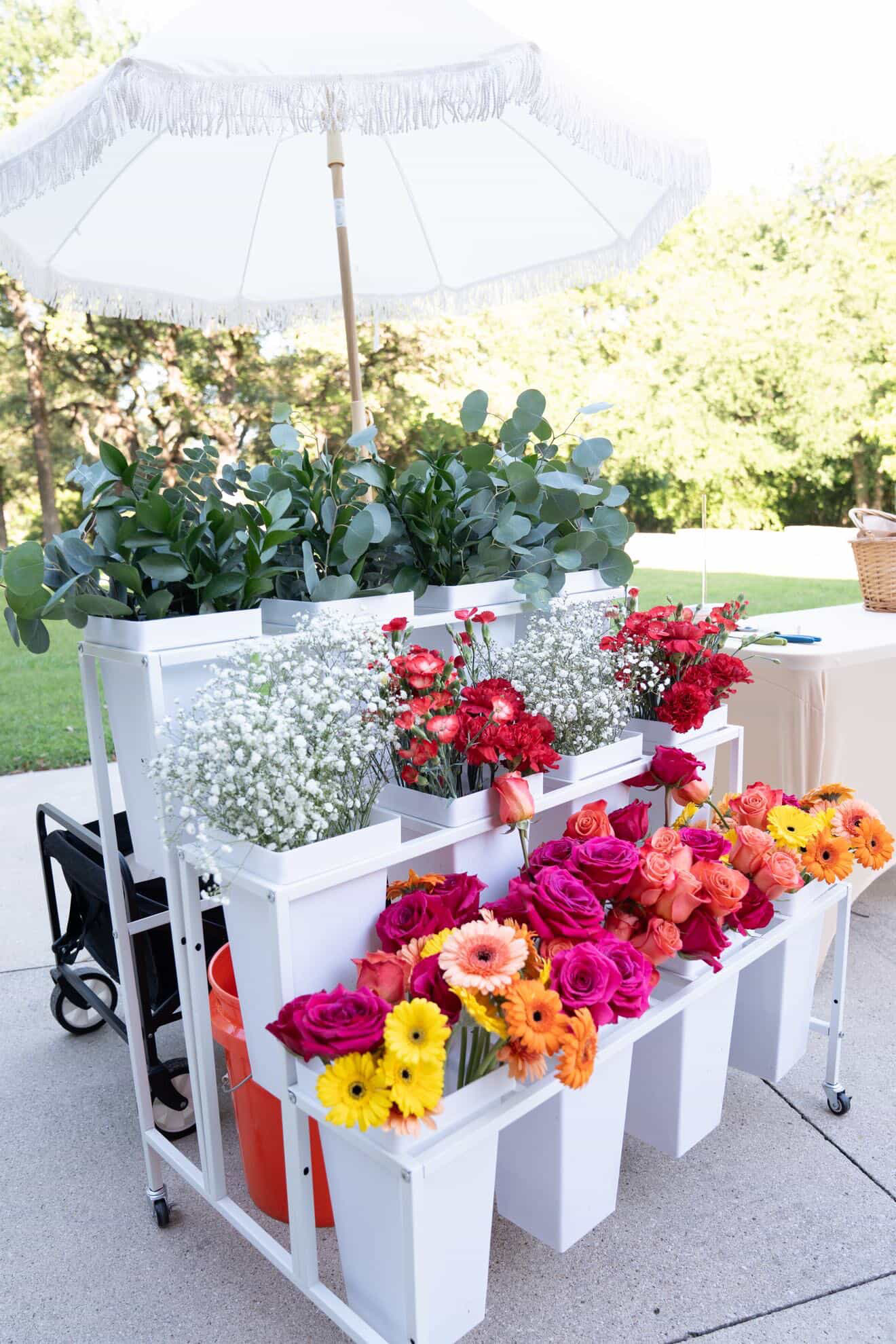 White flower stand with colorful flowers, umbrella, and greenery outdoors, bright sunlight, trees in background.